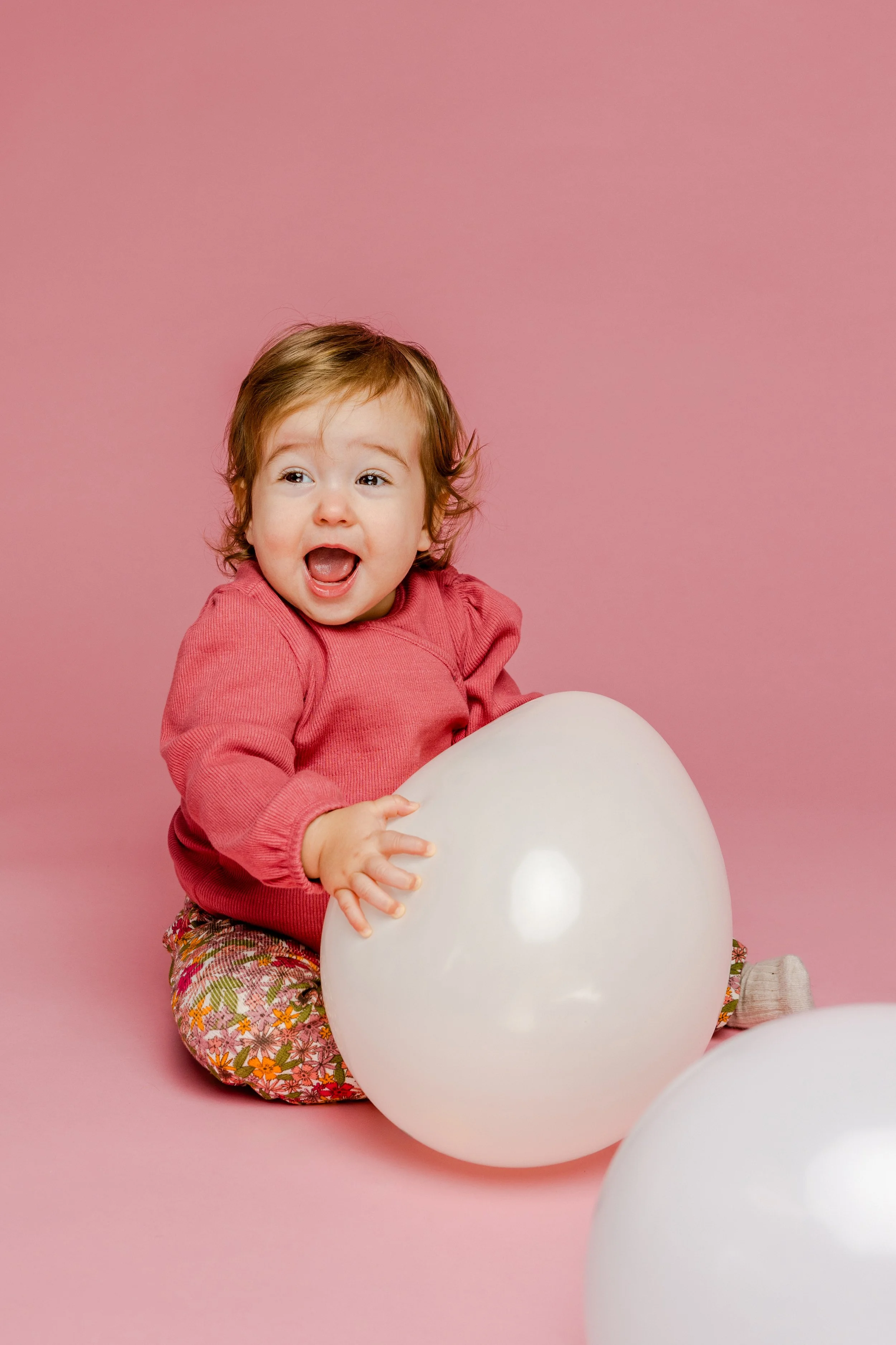 Een schattige jonge meid met bruin haar, gekleed in een roze top en bloemenbroek, zit op een roze achtergrond terwijl ze met witte ballonnen speelt en lacht.