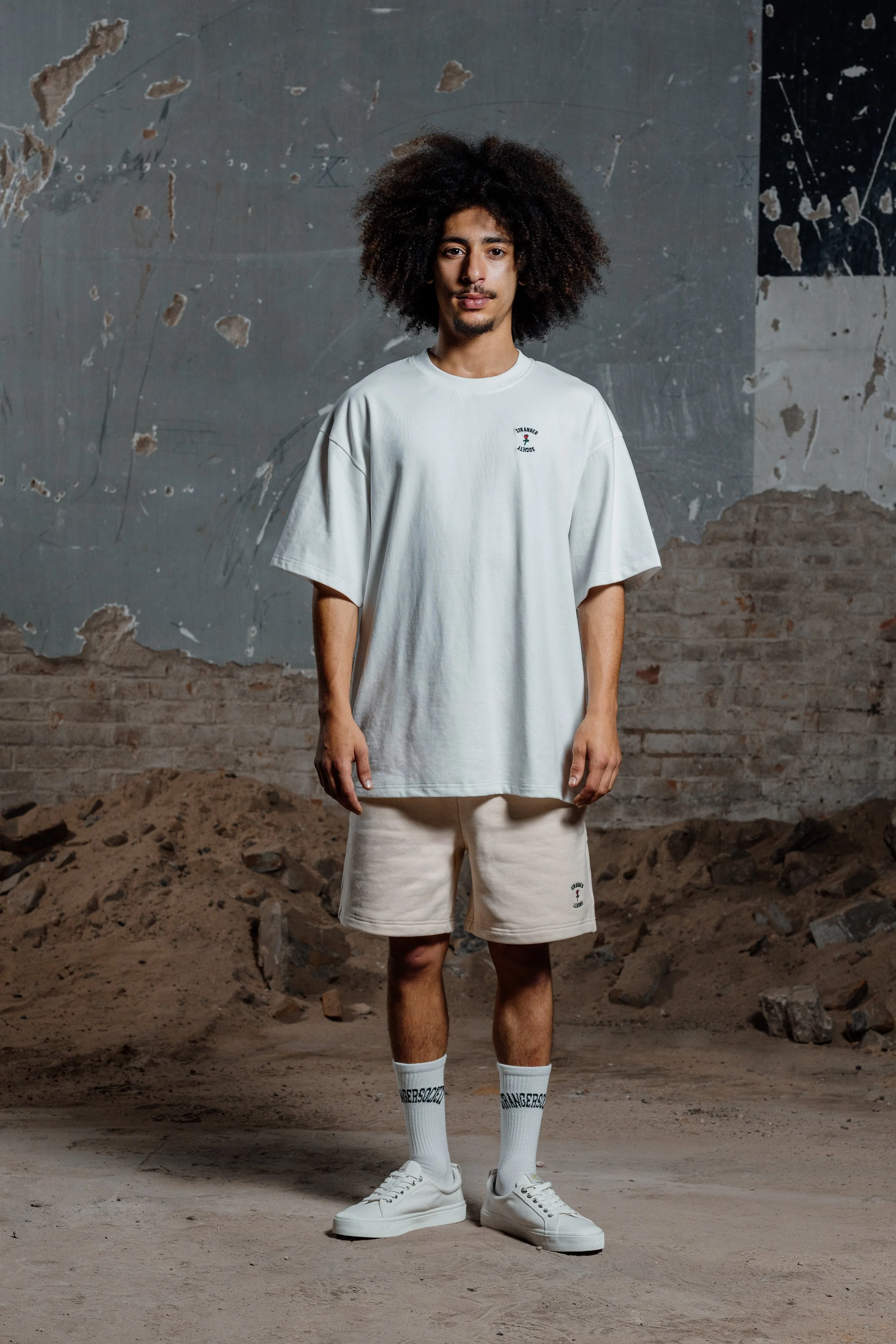 Een man in een witte oversized t-shirt, witte shorts, witte sokken en witte sneakers staat in een ruige, industriële omgeving met een gescheurde muur en een vloer van zand en stenen.