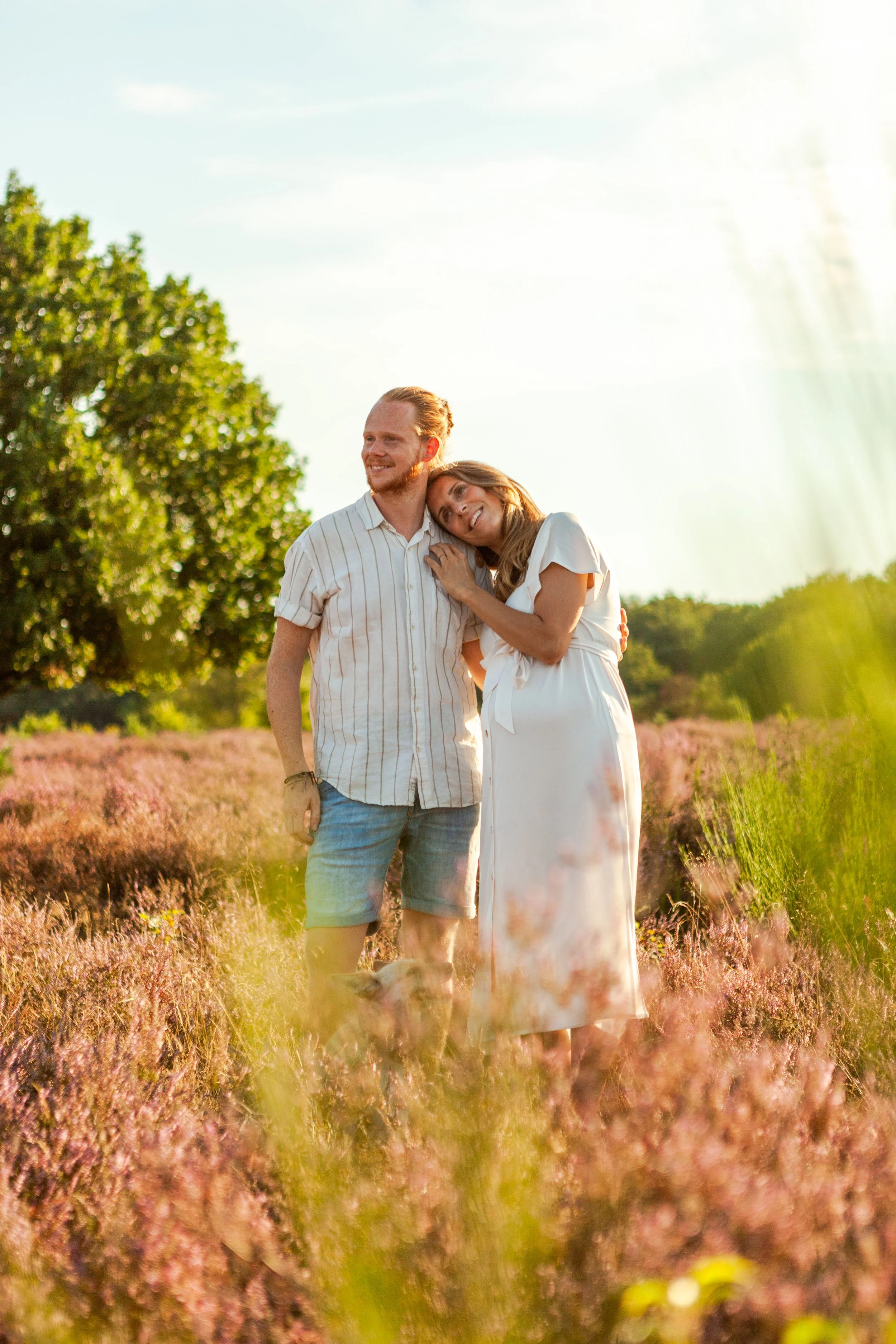 Een gelukkig paar staat samen in een veld met roze bloemen, omringd door groene bomen, tijdens zonsondergang.