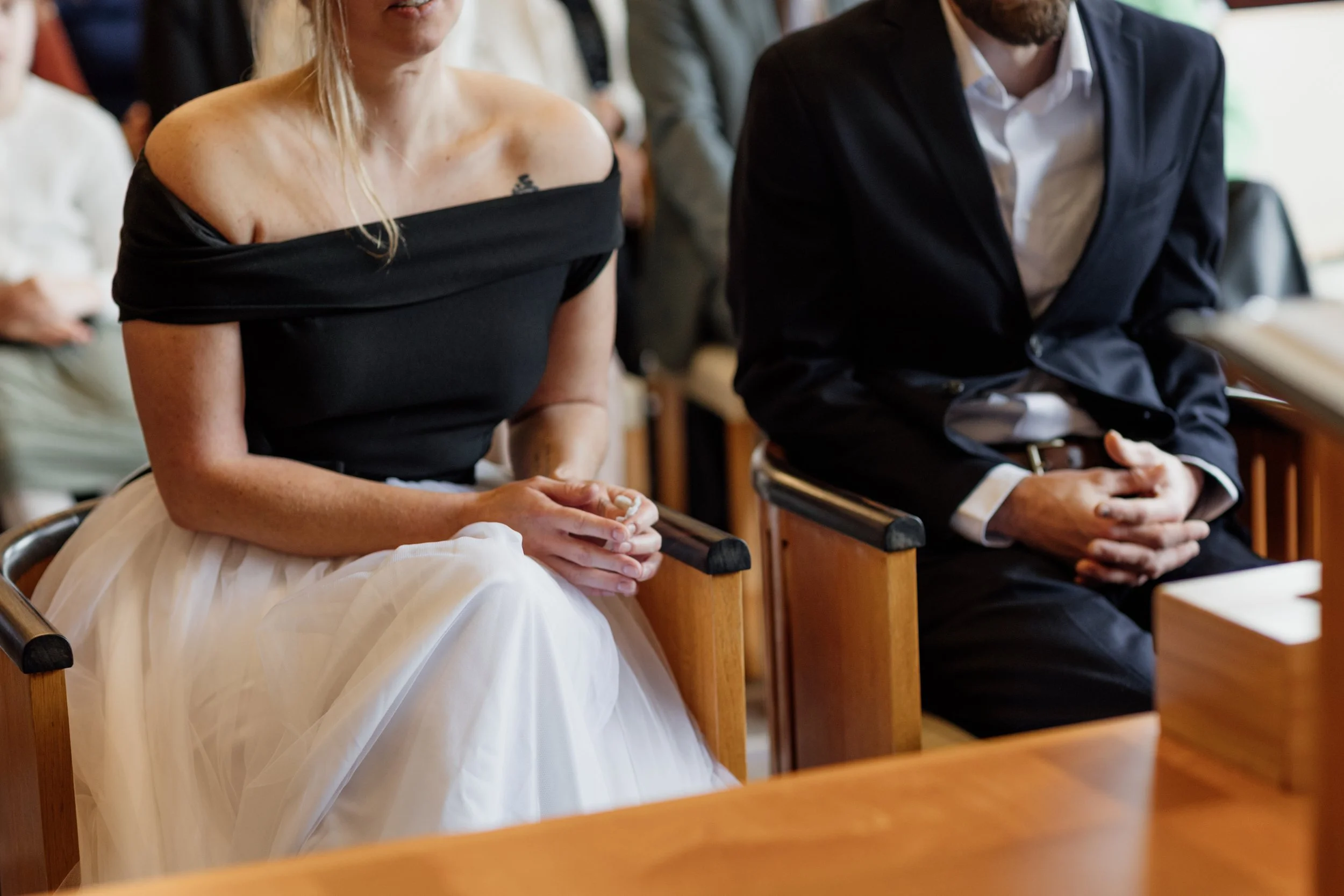 Twee mensen zitten in een kerk, betrokken bij een ceremonie. De vrouw draagt een off-shoulder zwart top en een witte rok, de man draagt een donkerblauw pak. Beiden hebben hun handen gevouwen.