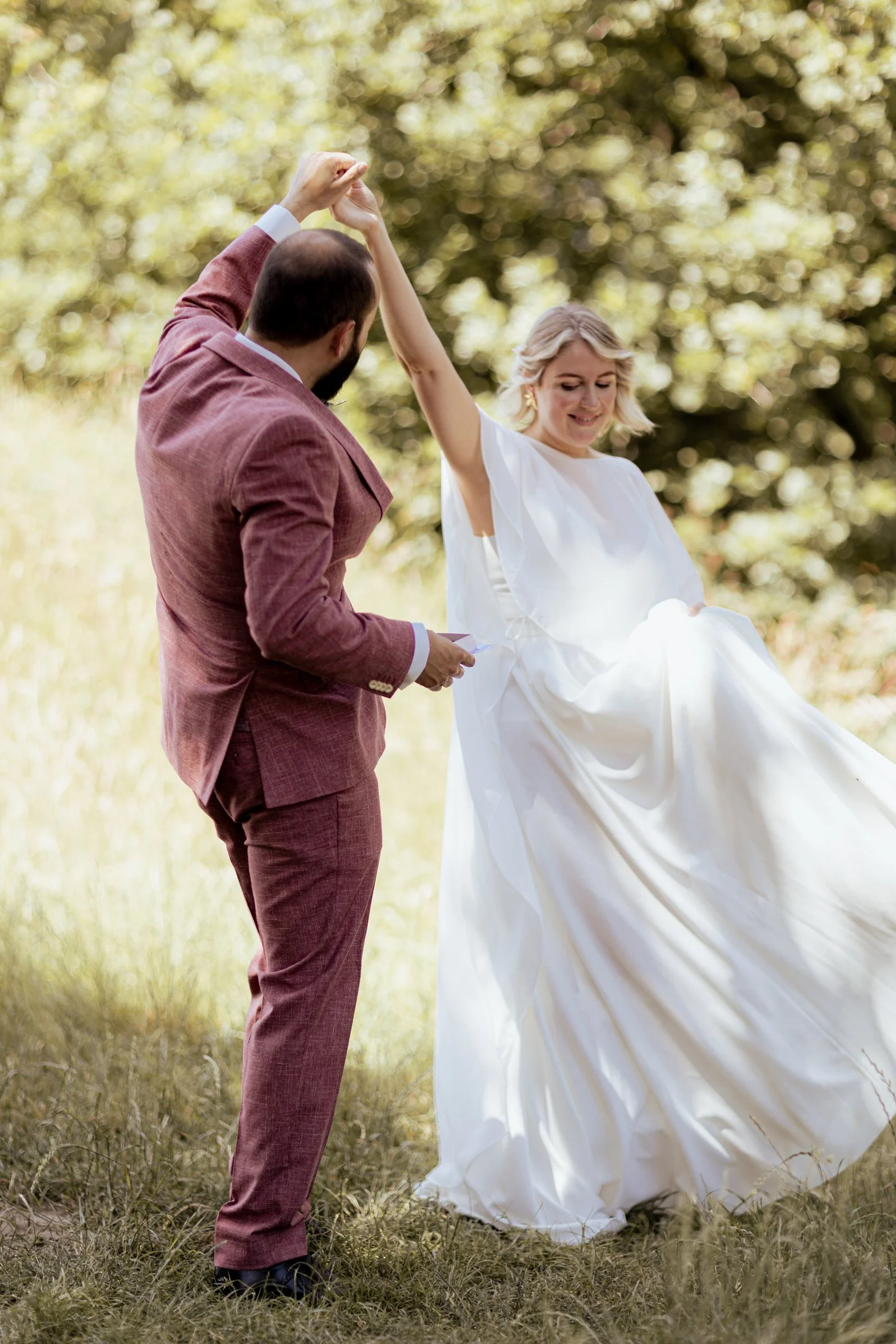 Een bruid en bruidegom dansen samen buiten in een park, omringd door groene bomen.