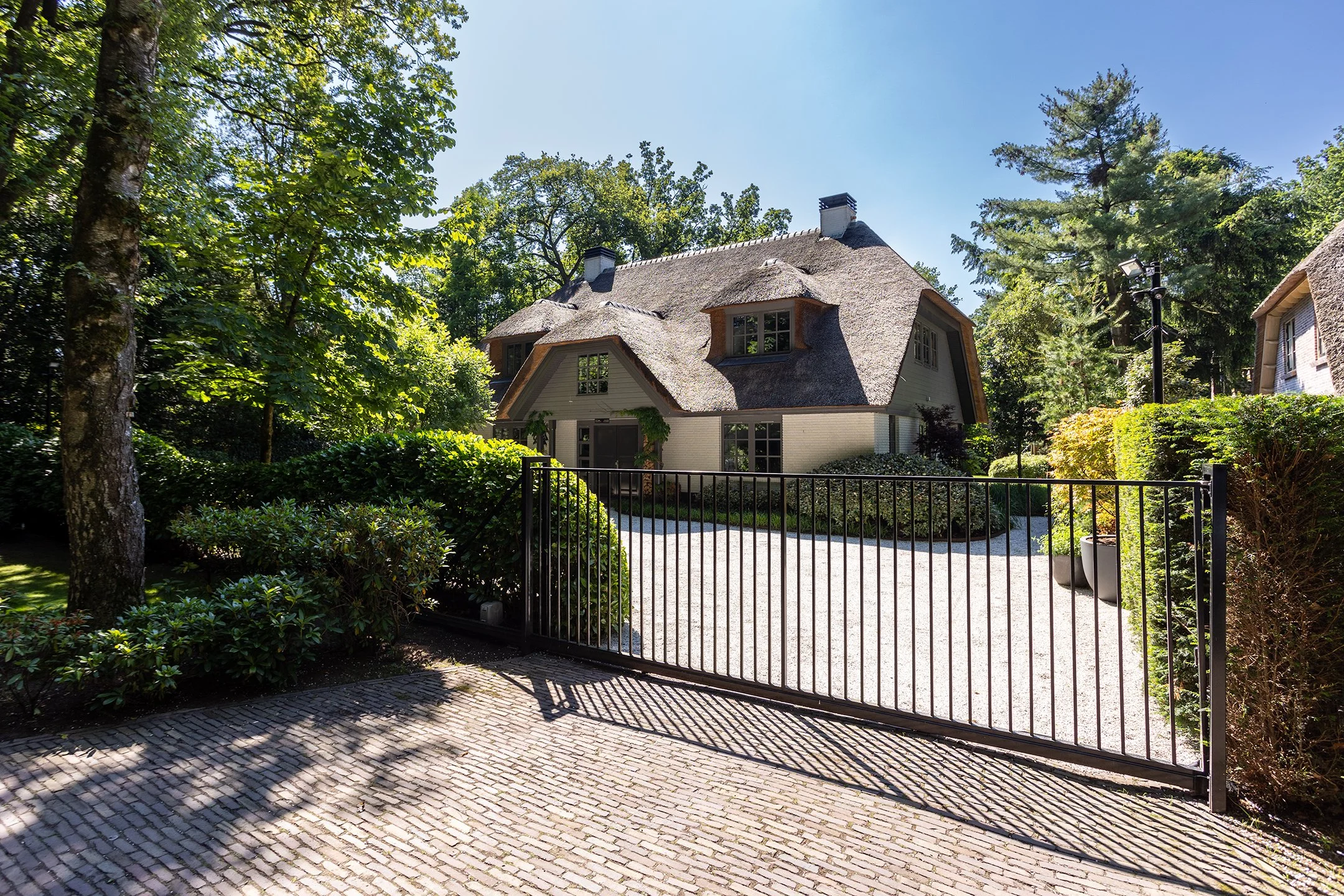 Een huis met rieten dak, omgeven door bomen en struiken, met een zwart metalen hek en een grindpad voor de ingang.