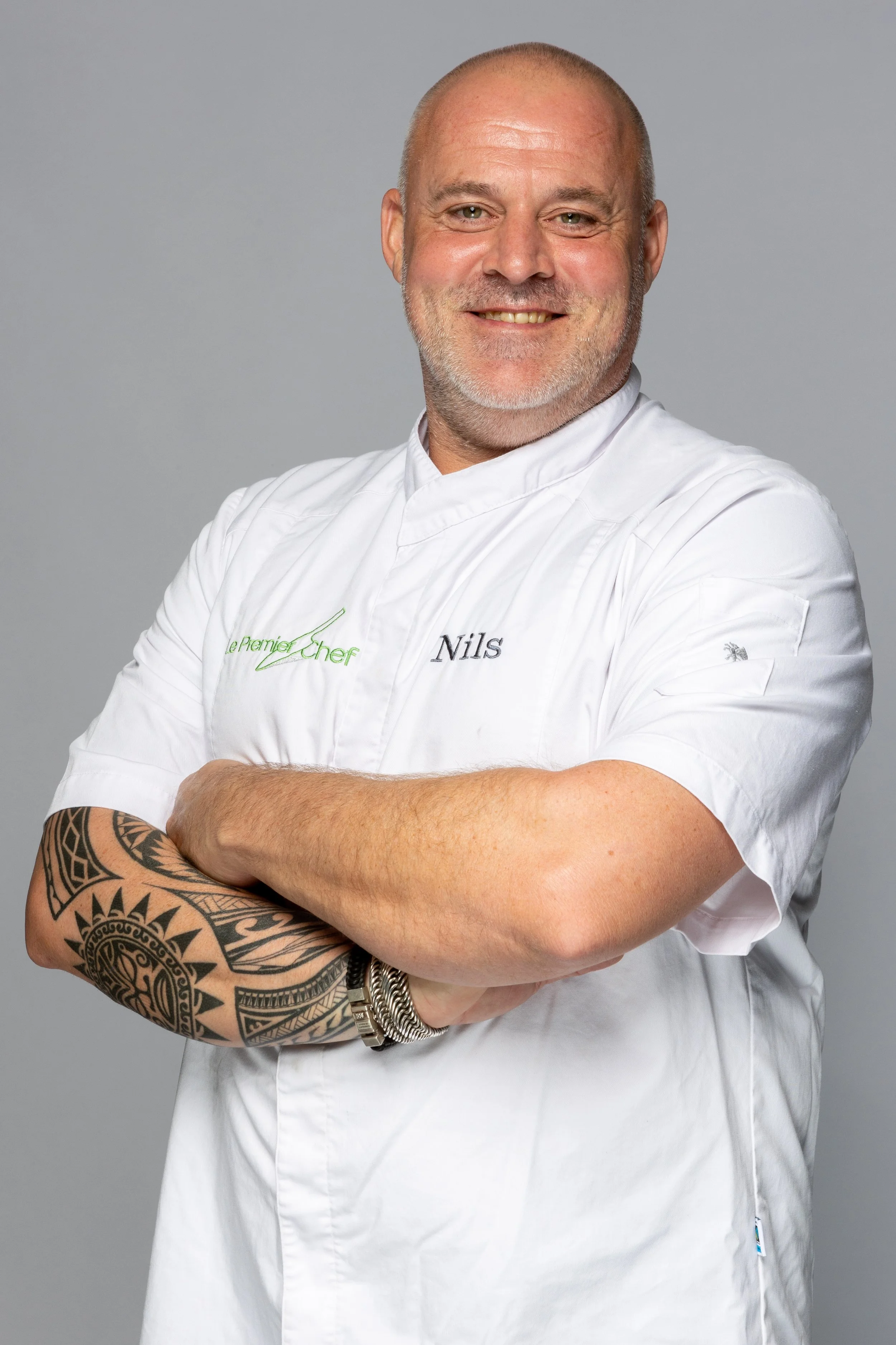 Persoon gekleed in een witte chef-kleding met de naam 'Nils', met een tattoo op de linker arm, poseert met gekruiste armen en lacht naar de camera, lichte achtergrond.