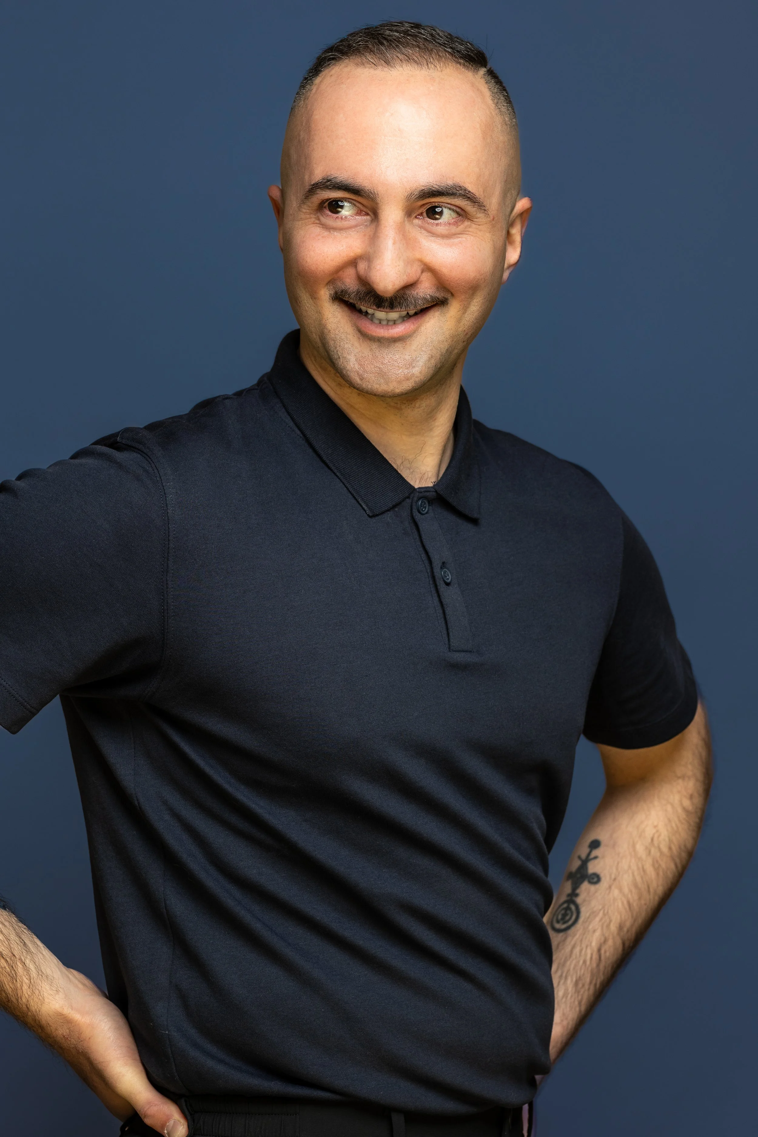 Portret van een lachende man met een getatoeëerde arm, gekleed in een zwarte polo, tegen een effen blauwe achtergrond.