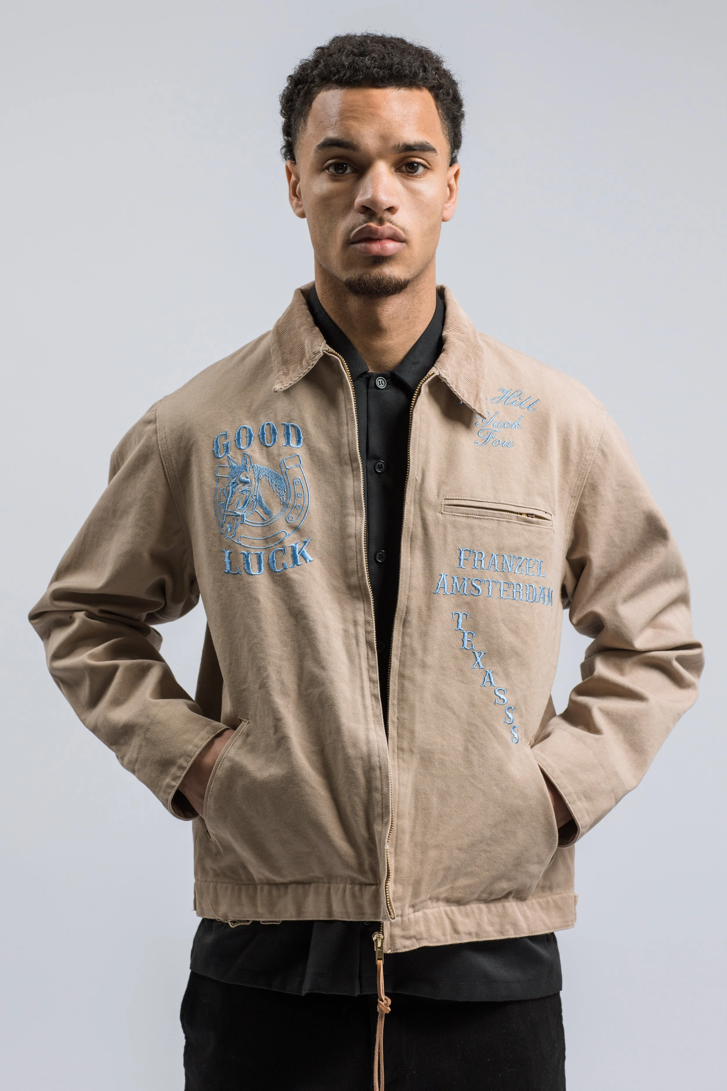 Jeune homme posant, portant une veste beige avec des motifs en filigrane bleus et des écritures, fond gris clair.