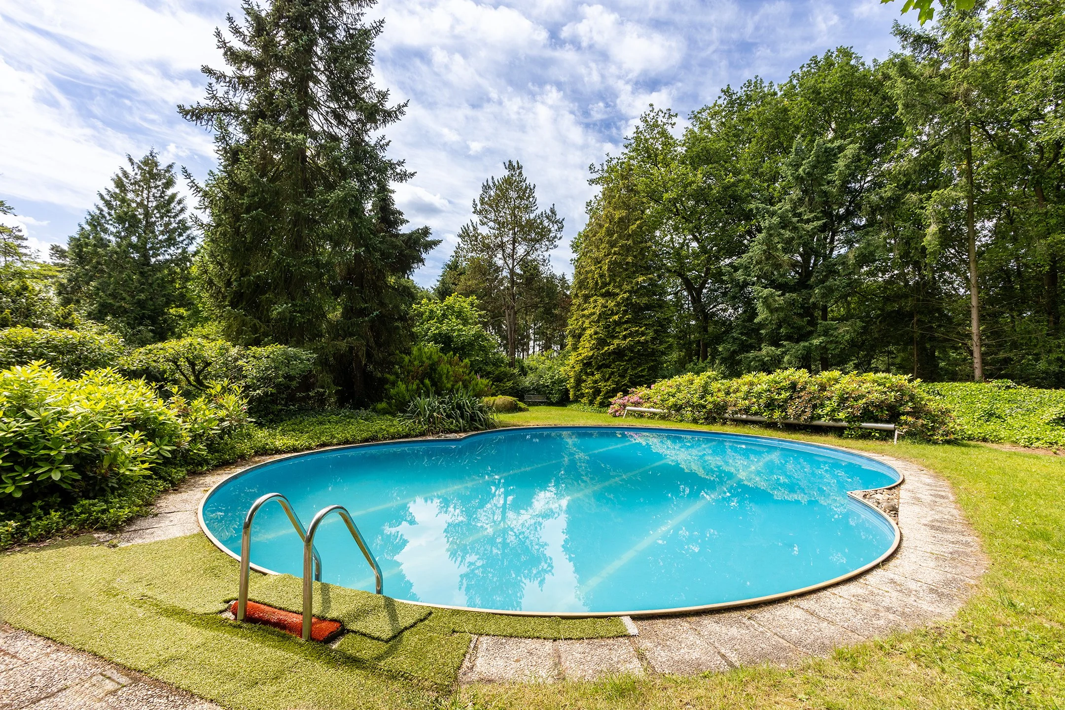 Ronde blauwe zwembad in een tuin met groene bomen en struiken onder een partly cloudy blauwe lucht.