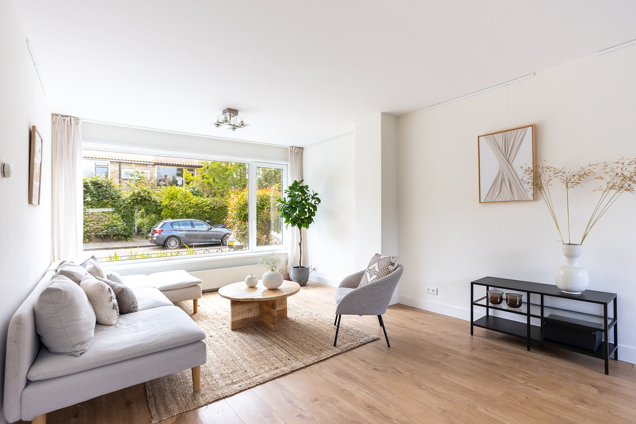 Minimalistisch ingerichte woonkamer met grote raam, witte bank, grijze fauteuil, planten, houten vloer, beige tapijt, en zwart tv-meubel met witte vaas en decoraties.