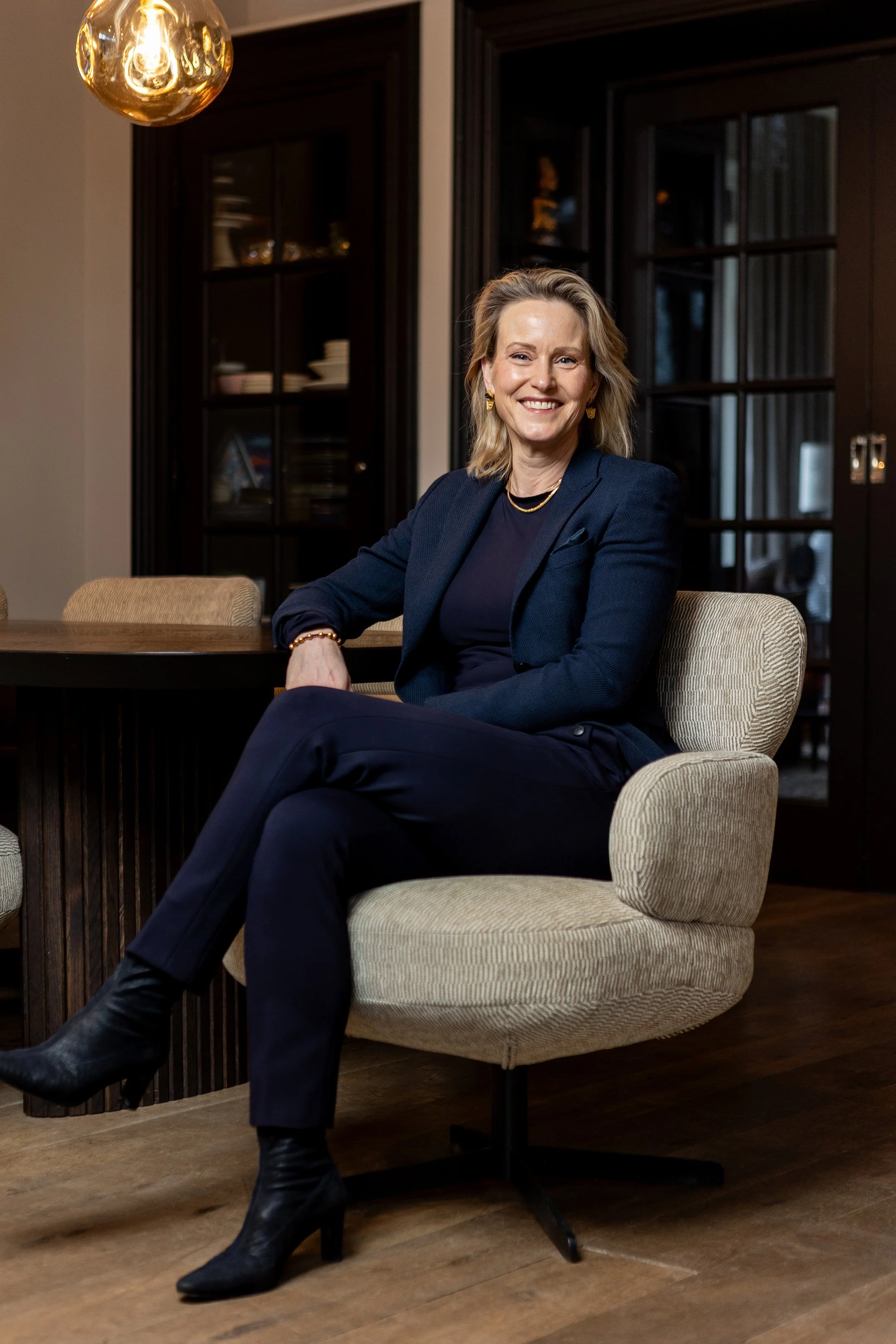 Een vrouw in een donkere blazer en broek zit op een beige stoel in een interieur met een donkere houten kast en glazen deuren, lachend naar de camera.