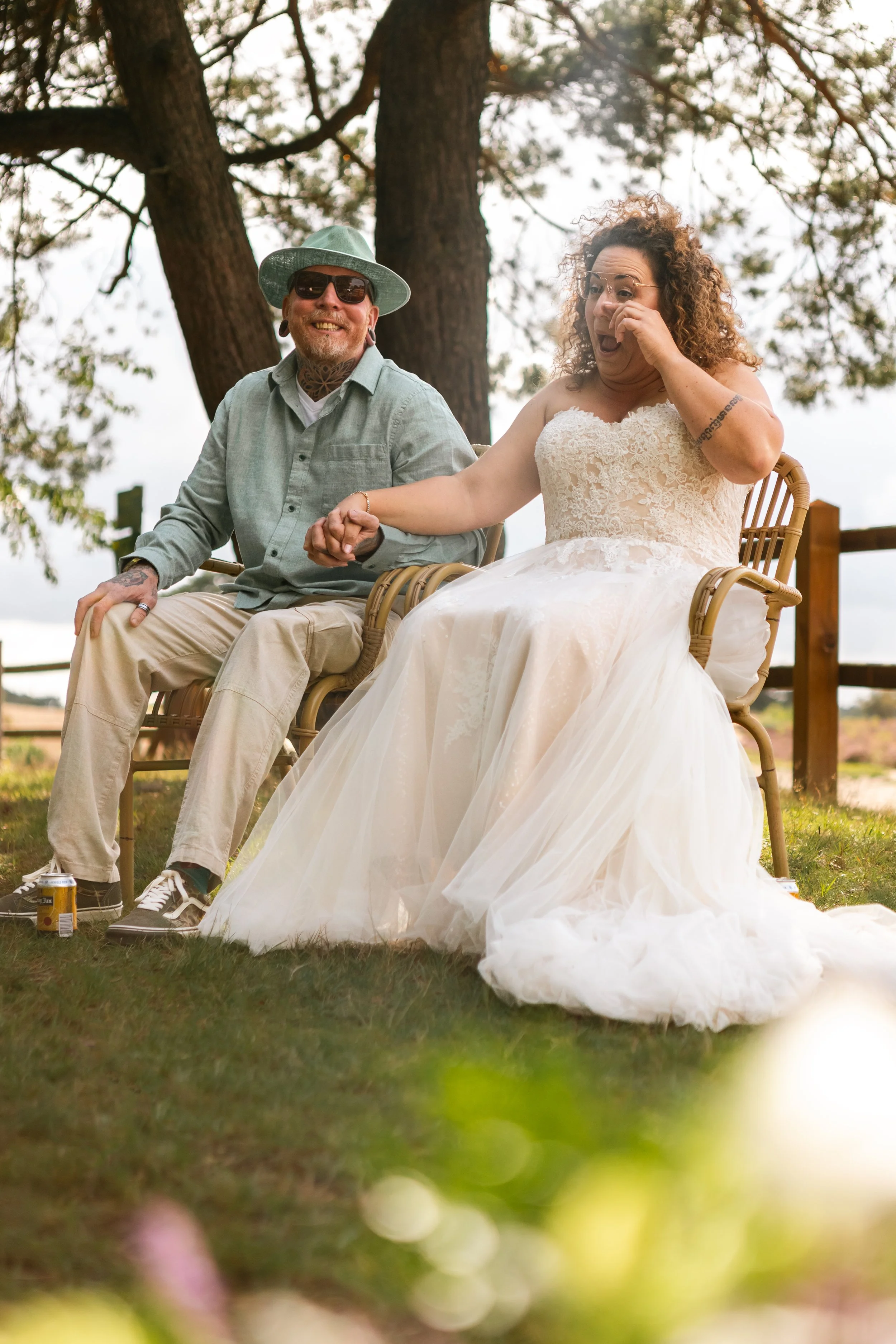 Twee mensen, een man en een vrouw in een witte trouwjurk, zitten op houten stoelen buiten onder een boom. Ze lijken op een bruiloft en ze lachen en hebben plezier.