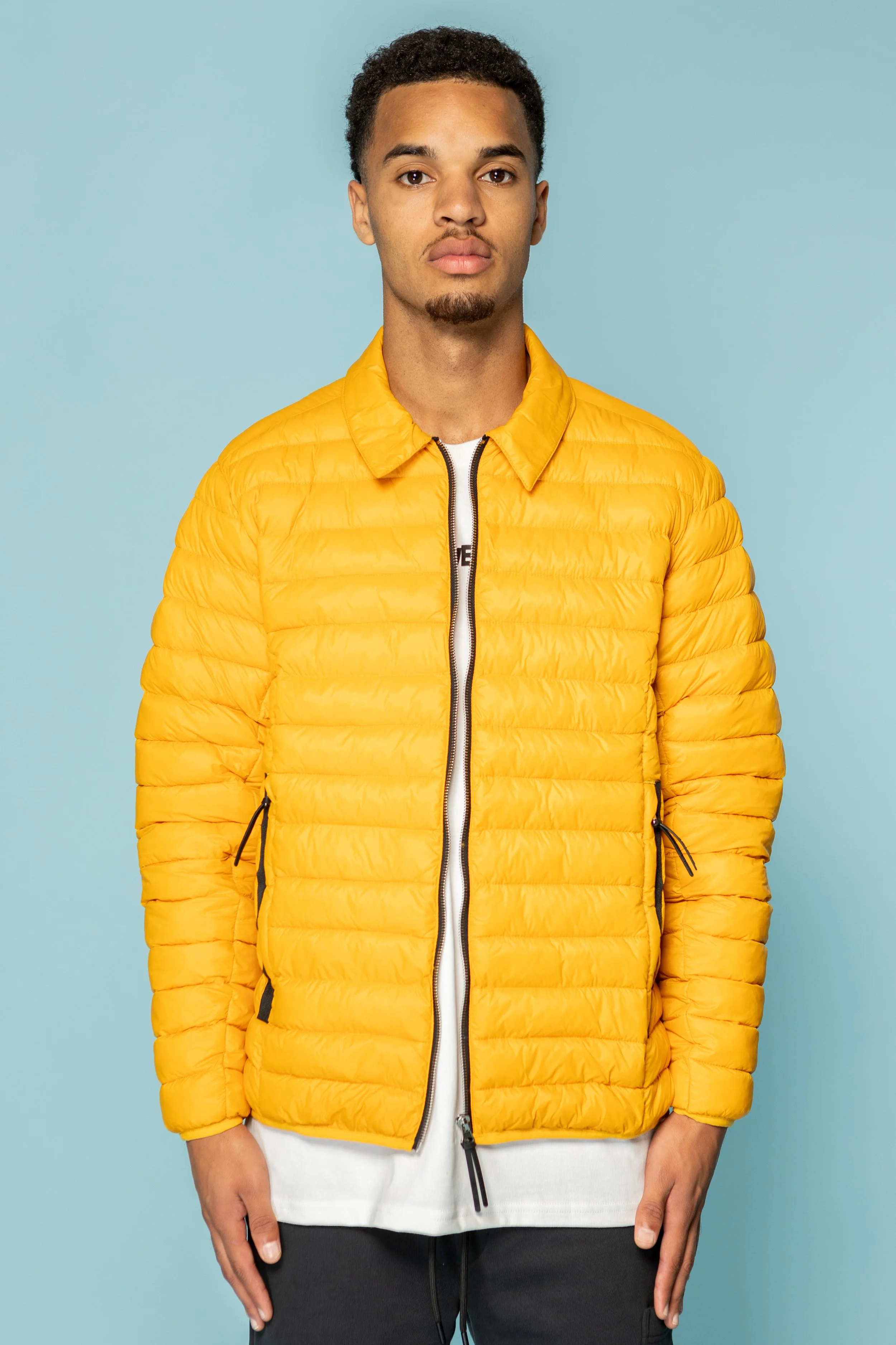 Man in een gele pufferjas met zwarte ritssluiting, zwarte details, en witte kleding eronder, staand voor een lichtblauwe achtergrond.