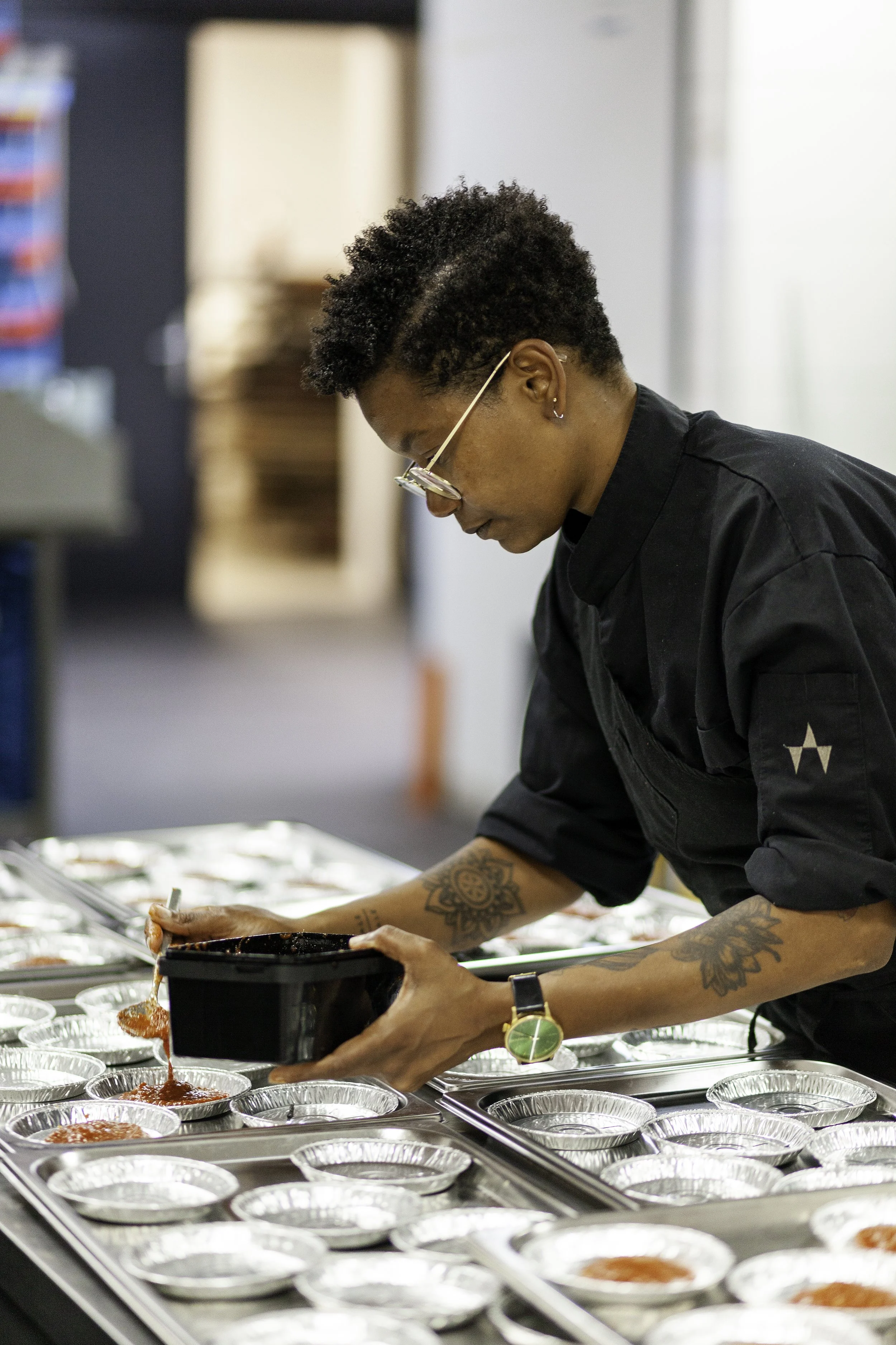 Een chef-kok met korte, gekrulde haar, oorbel en tattoos op de armen, schept tomatensaus in kleine aluminium bakjes in een keukenrestaurant.