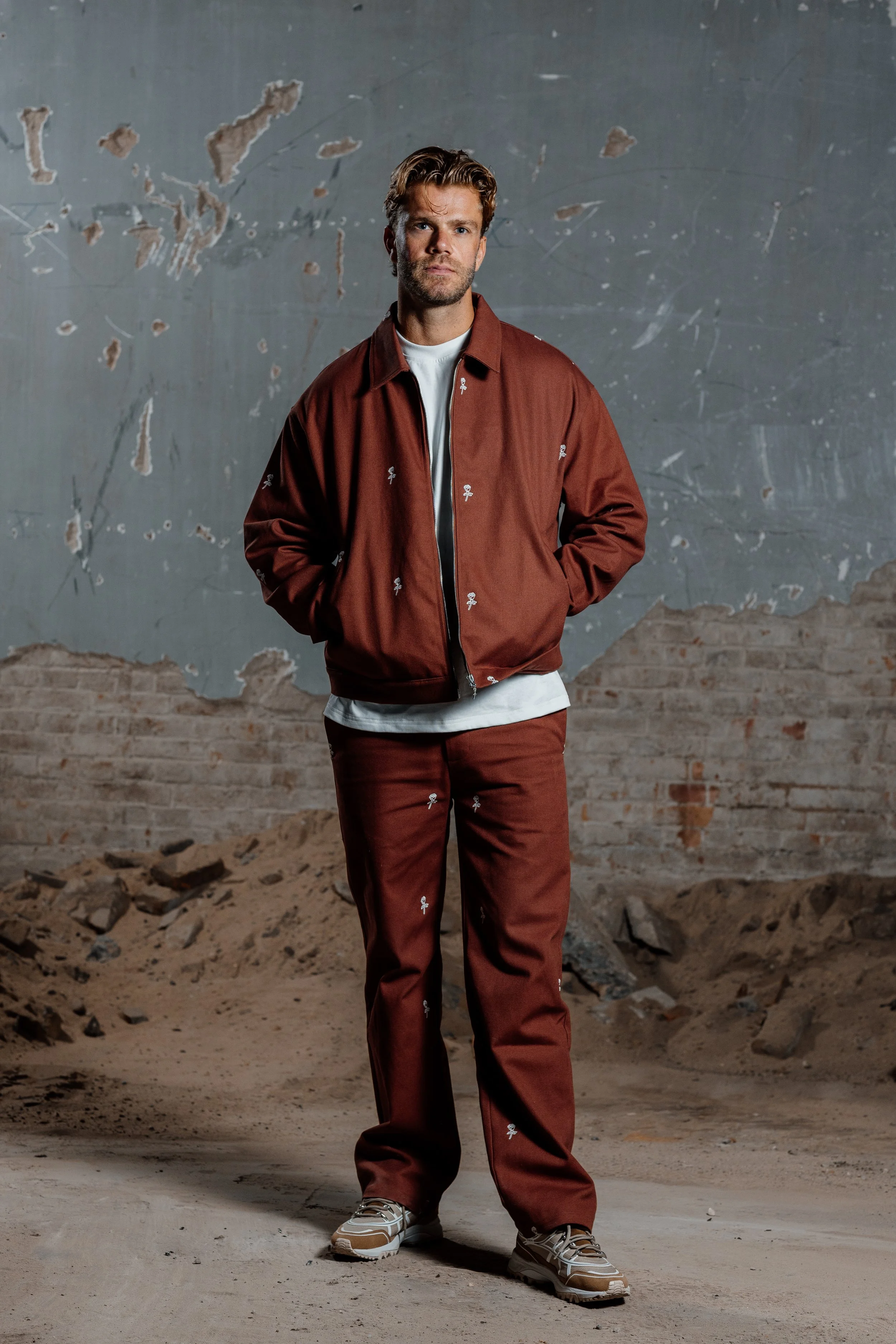 Man poseert in een industrieel decor met oude muur en zand op de grond, gekleed in een bruin trainingsjack en broek met een wit shirt eronder, sneakers.
