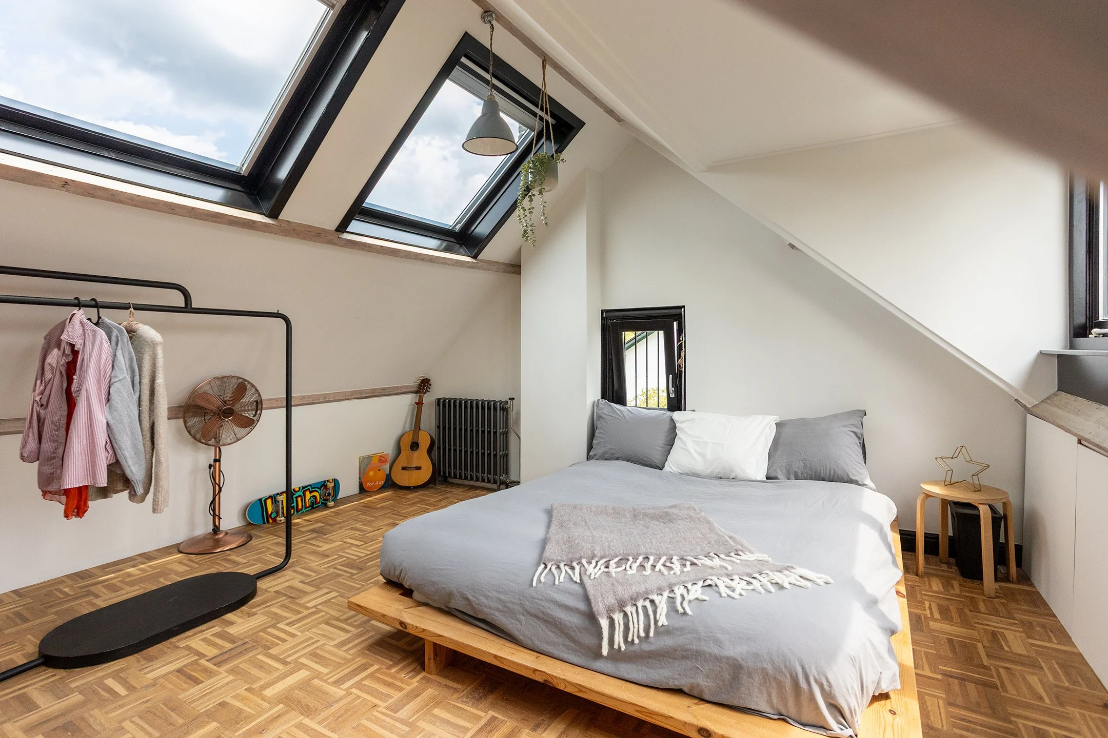 Een slaapkamer met een bed, twee kussens, een deken, een kleine zwart-grijze nachtkast, een kamerscherm met een kledingrek, een ventilator, een gitaar, een skateboard, en een raam onder een schuine dak. Een plafondlamp en een klein plafondraam zorgen