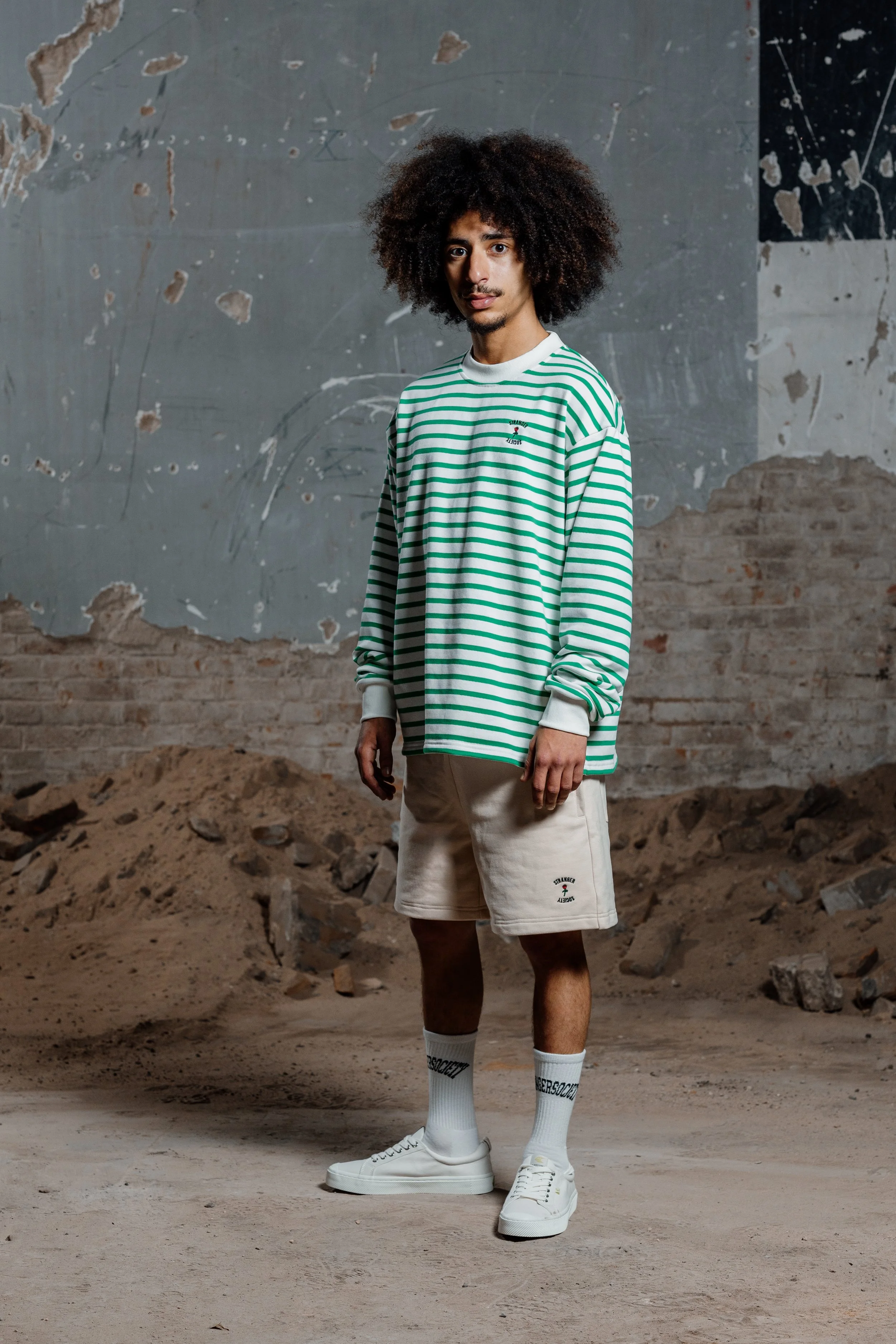 Man draagt een groene en witte gestreepte trui en beige korte broek, met witte sneakers en witte sokken, in een ruïne-achtige omgeving.