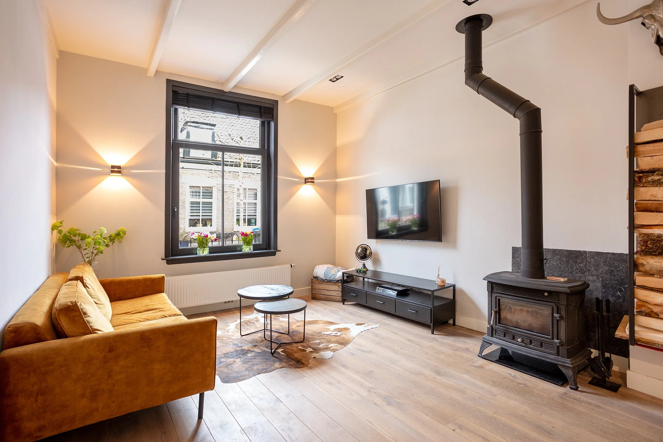 Woonkamer met gele bank, ronde tafels, muur met tv, haard en houtopslag, raam met bloemen, houten vloeren en muurverlichting.