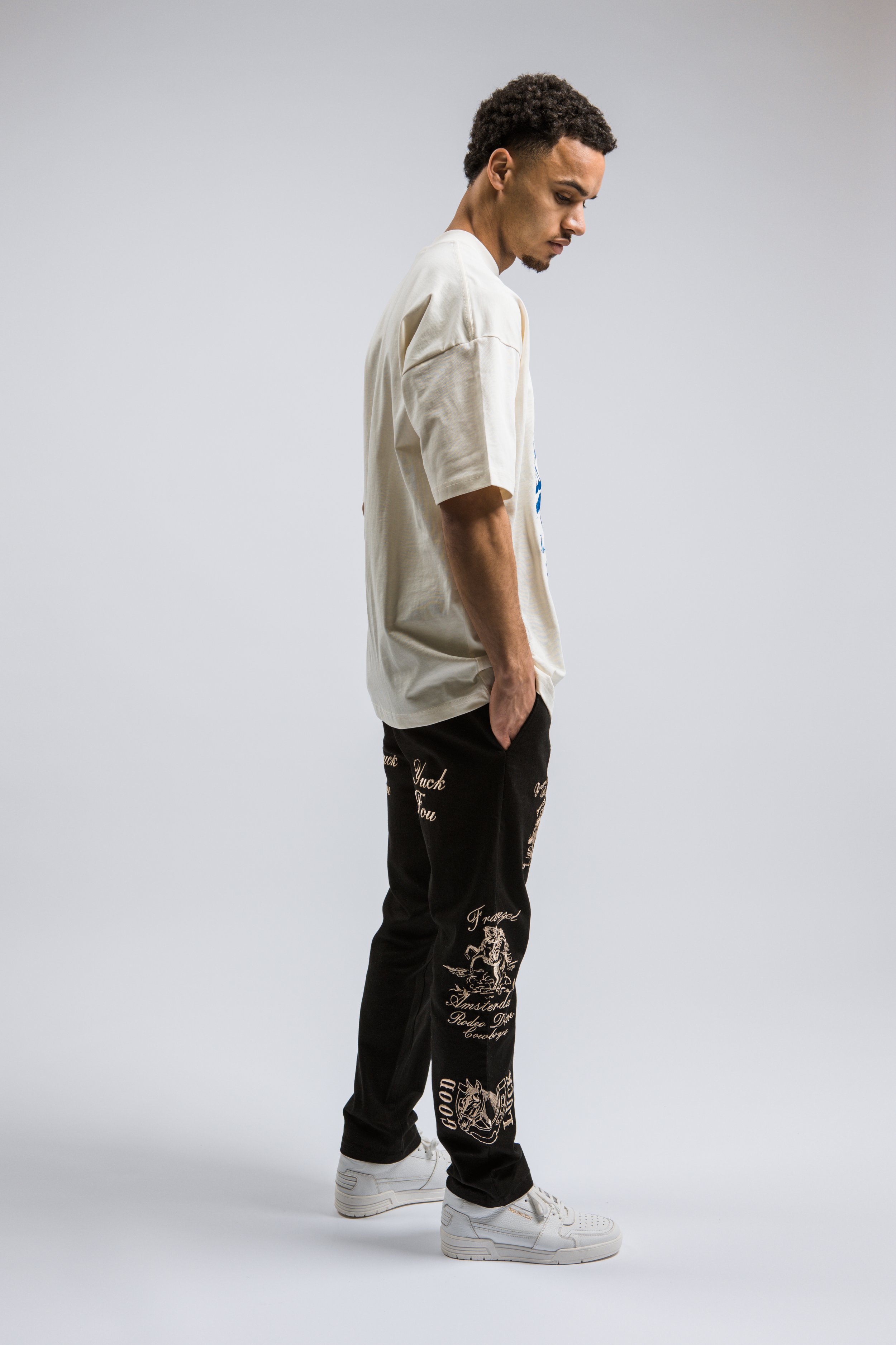 Een jonge man staat op een witte achtergrond, gekleed in een beige T-shirt, zwarte joggingsbroek met teksten en afbeeldingen, en witte sneakers.