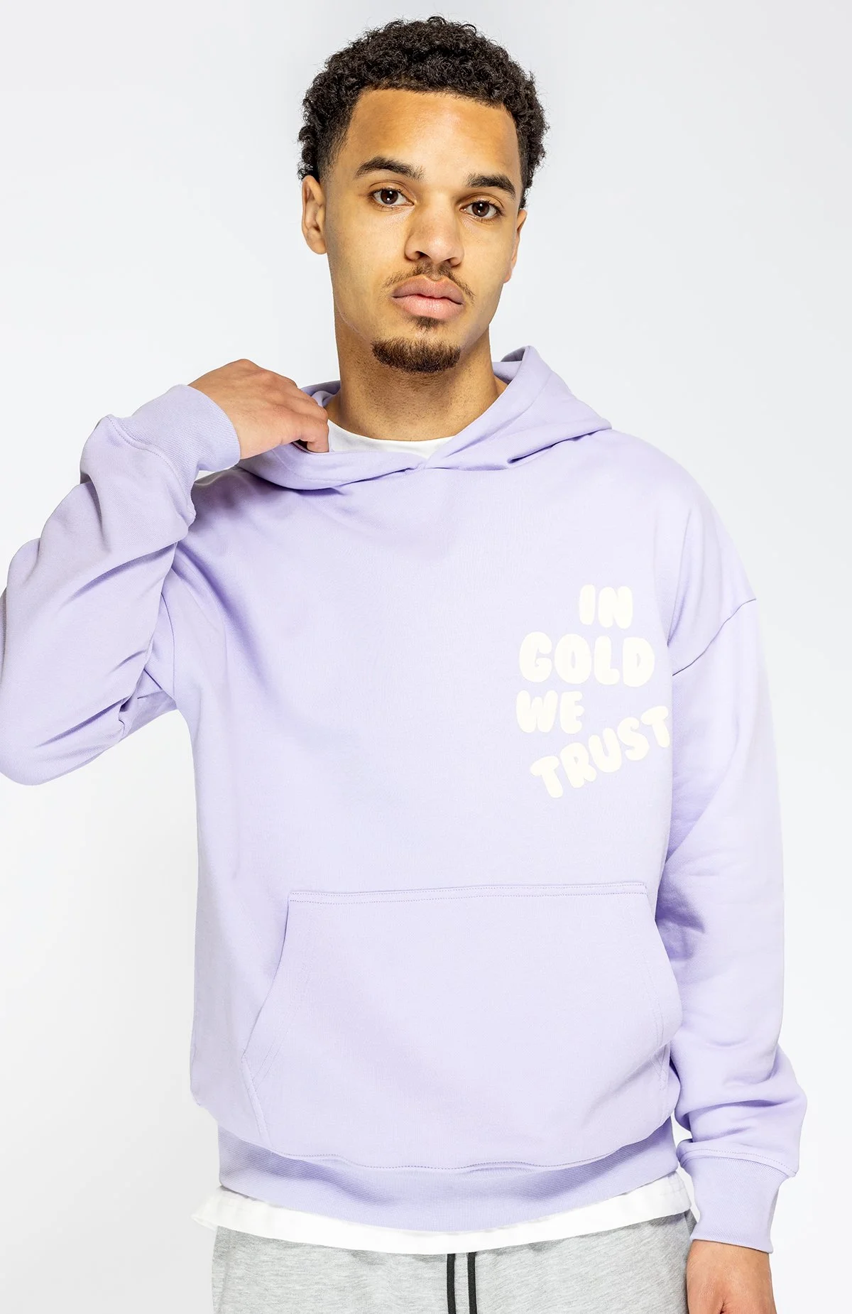 Man die een lichtpaarse hoodie draagt met de tekst 'In Gold We Trust' op de voorkant, tegen een witte achtergrond. Proxy met een hand die de hoodie aan de hals vastpakt.