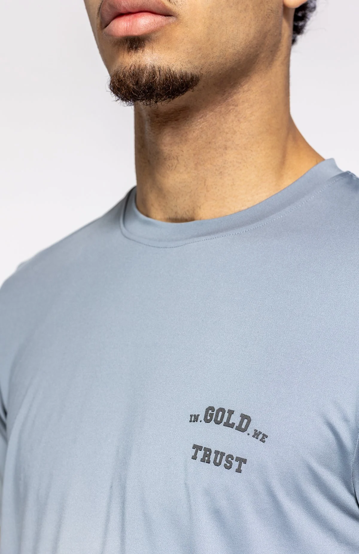 Close-up van een man met een kort gezichtshaar en een lichte blauwe T-shirt met de woorden 'IN. GOL.D' en 'TRUST' erop.