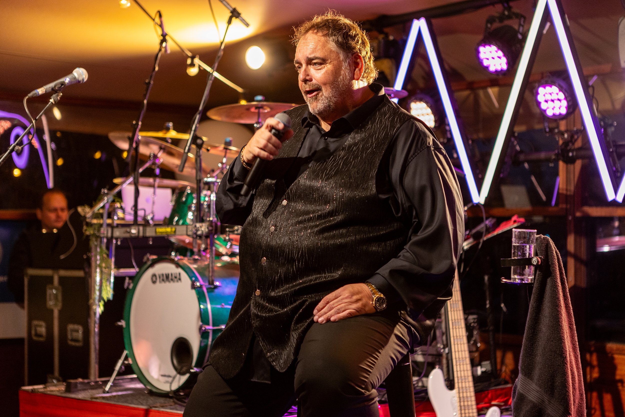 Man zit op een stoel en houdt een microfoon vast, terwijl hij op het podium staat met muziekinstrumenten en lampen op de achtergrond.