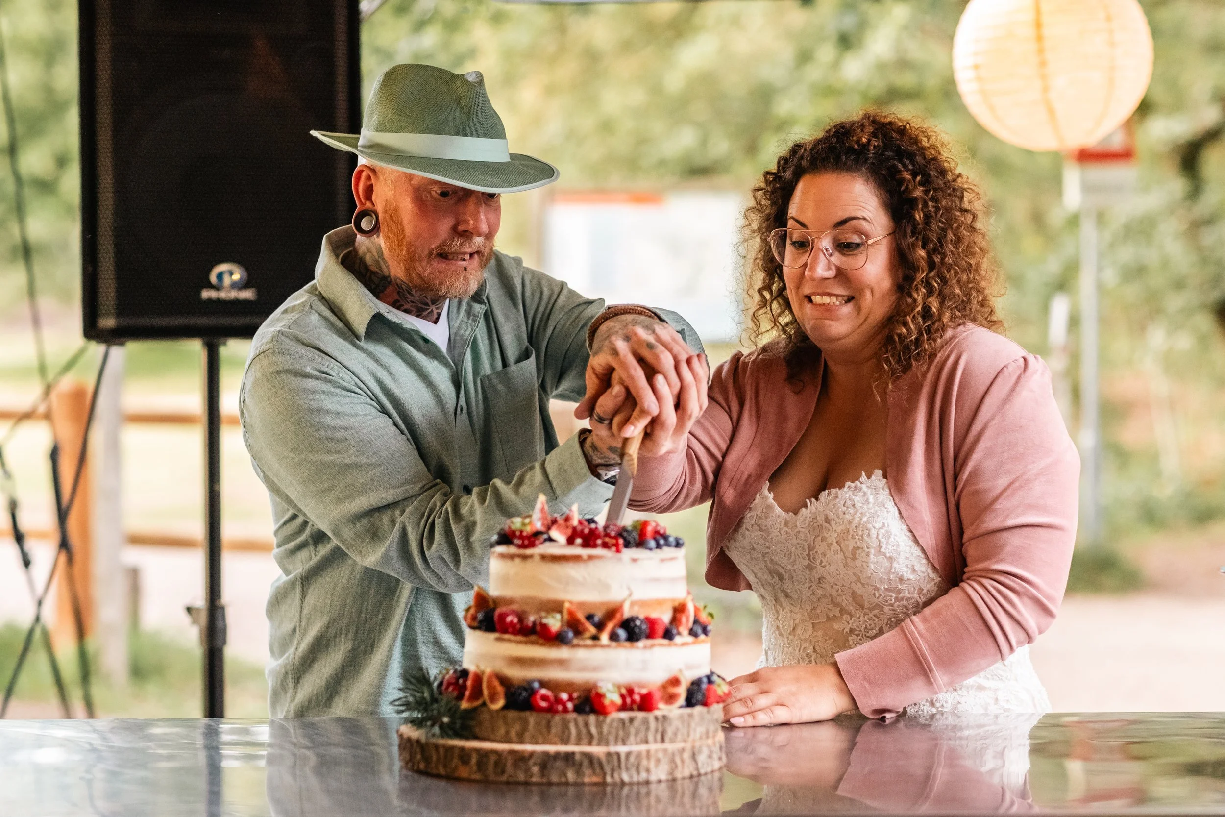 Man en vrouw snijden samen in een feesttaart met bessen en vruchten tijdens een viering, binnen een feestzaal met lampionnen.