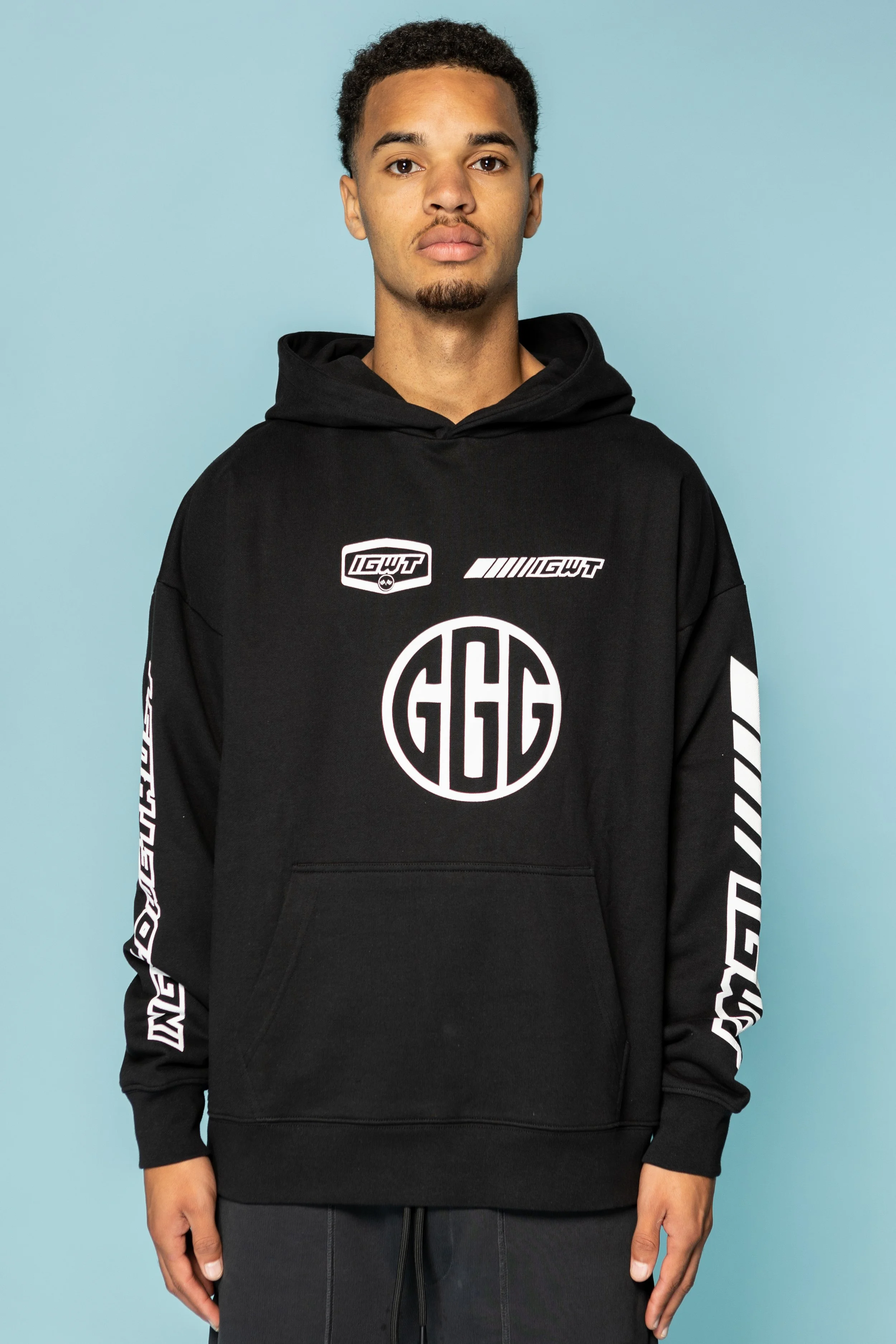Een jonge man draagt een zwarte hoodie met witte grafische teksten en logo's, voor een lichtblauwe achtergrond.