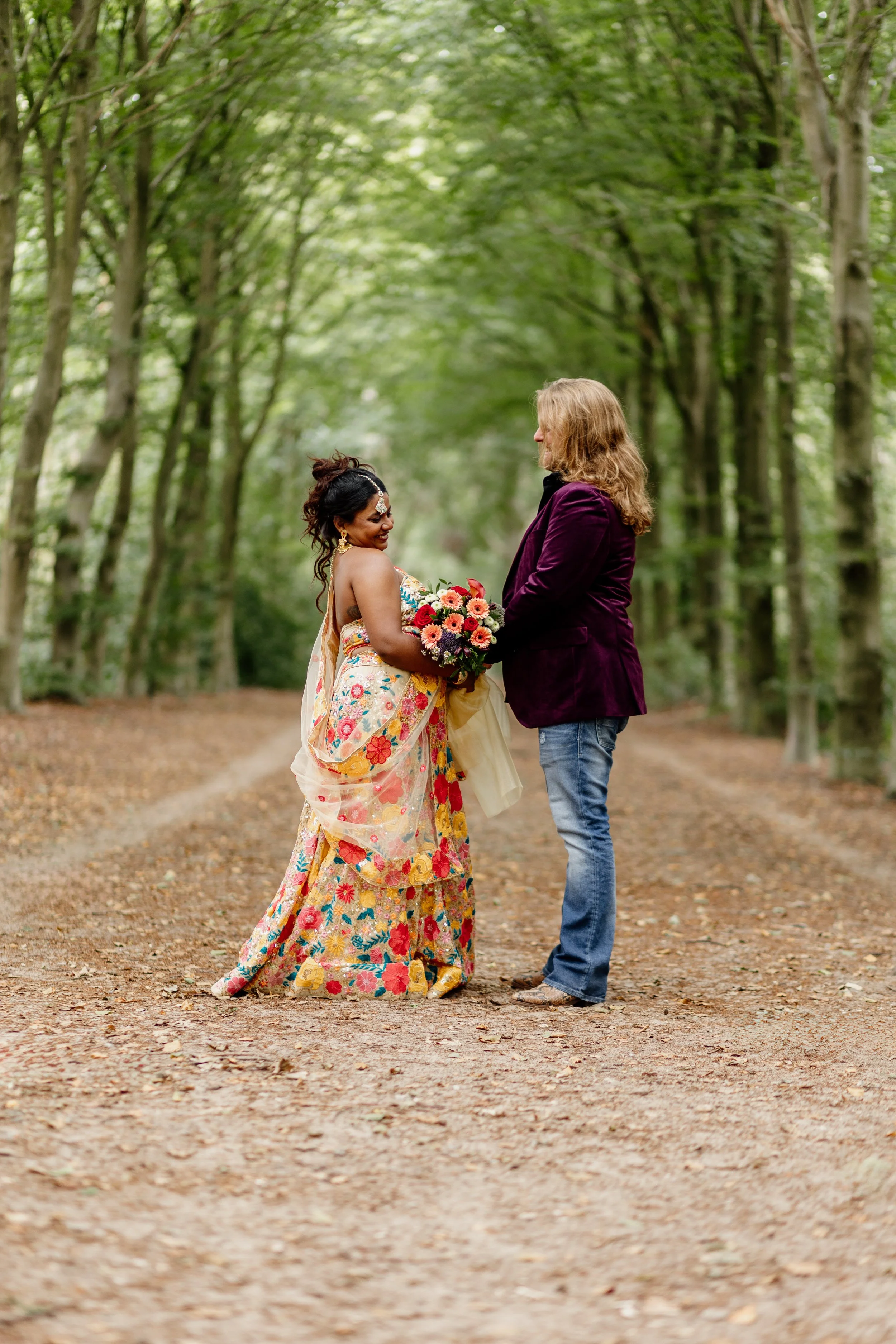 Twee mensen staan op een bosweg, een vrouw in een kleurrijk feestlijk jurk met bloemen en een man in een paarse jas, die elkaar de hand vasthouden.