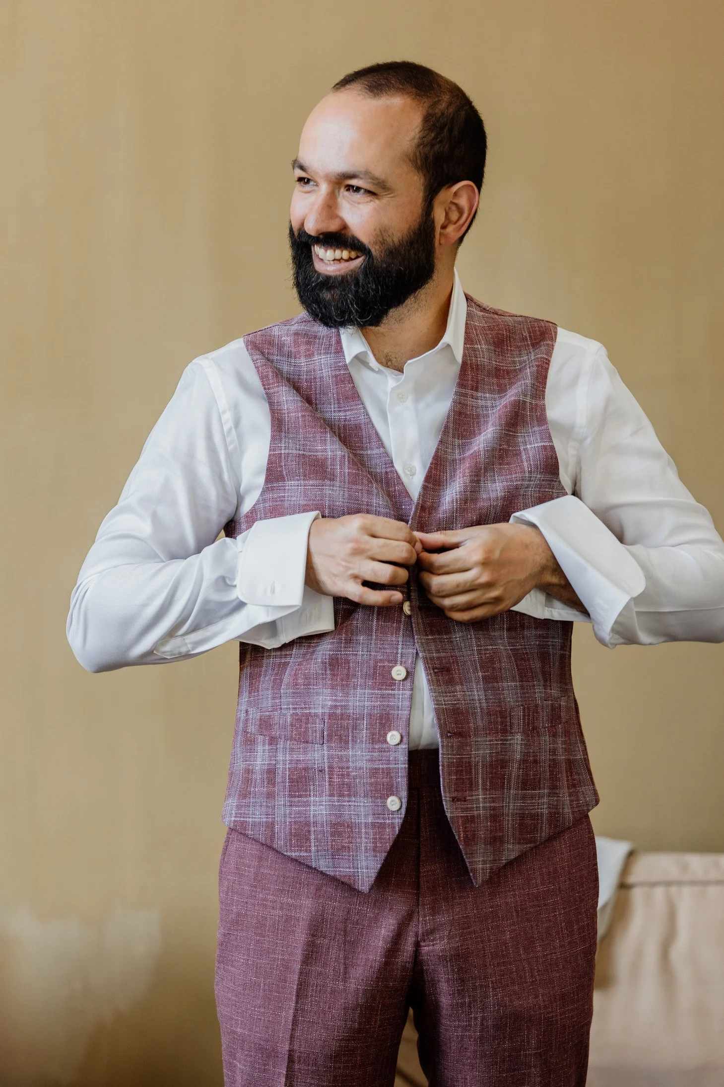 Man met een volle baard en kort haar, die een wit overhemd en een rood geruite vest en broek draagt. Hij lacht en knoopt zijn vest dicht.