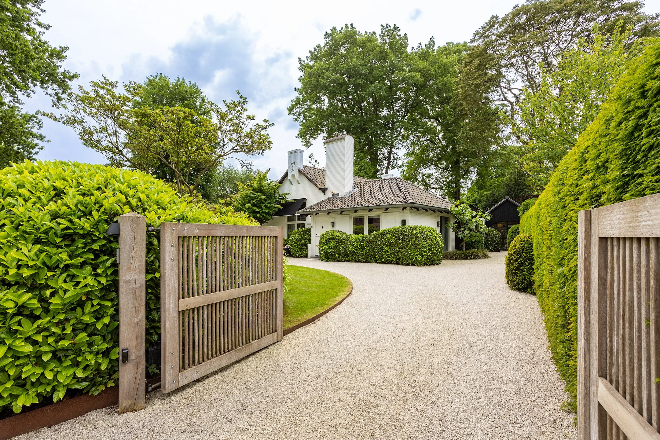 Een wit huis met een dak van tegels, omgeven door groene bomen en struiken, met een grindpad dat naar de ingang leidt, en een houten hek aan de ingang.