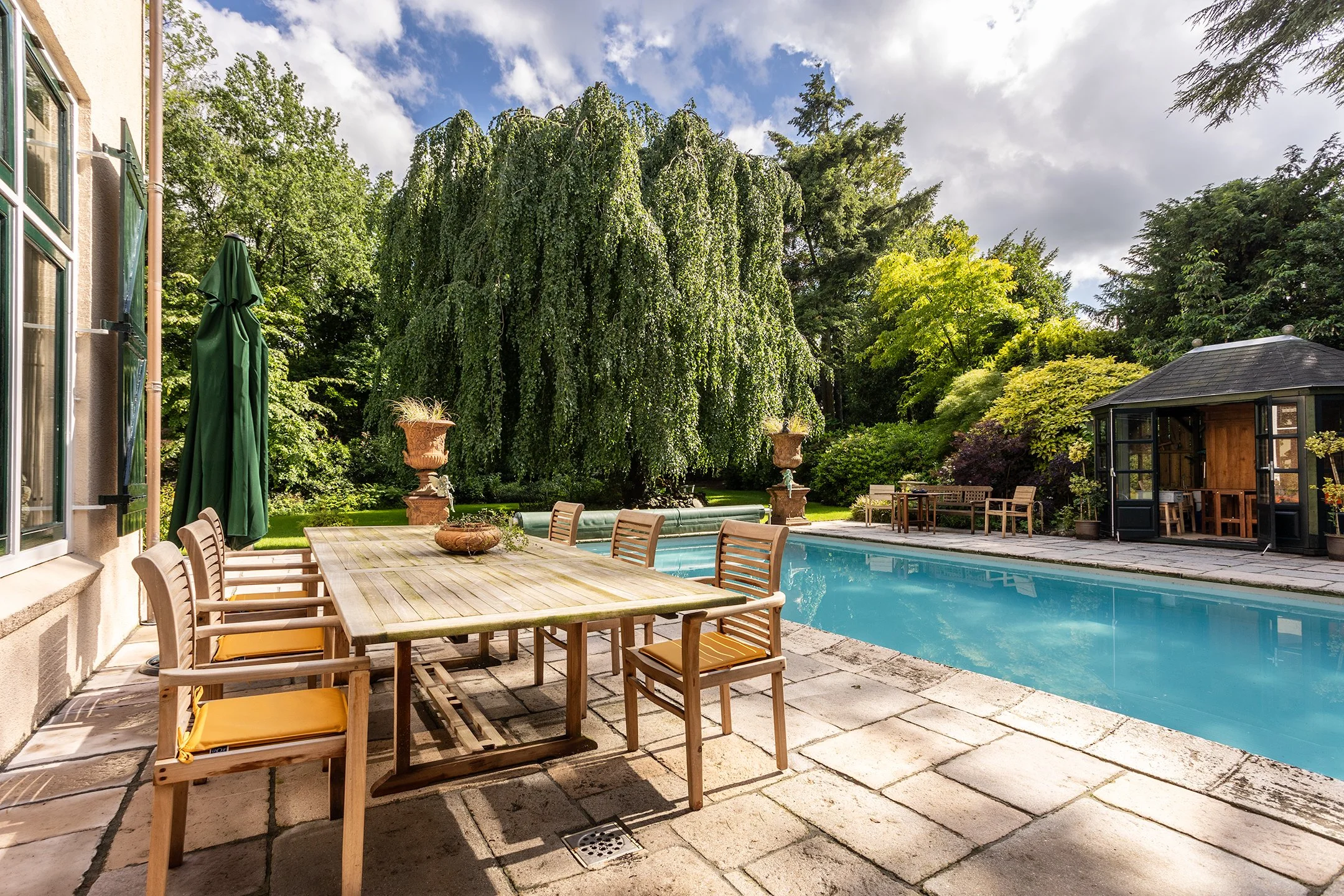 Uitzicht op een tuin met een zwembad, een houten tafel met stoelen, een groene parasol en grote plantenbakken. Er staan bomen, struiken en een gazebo op de achtergrond.