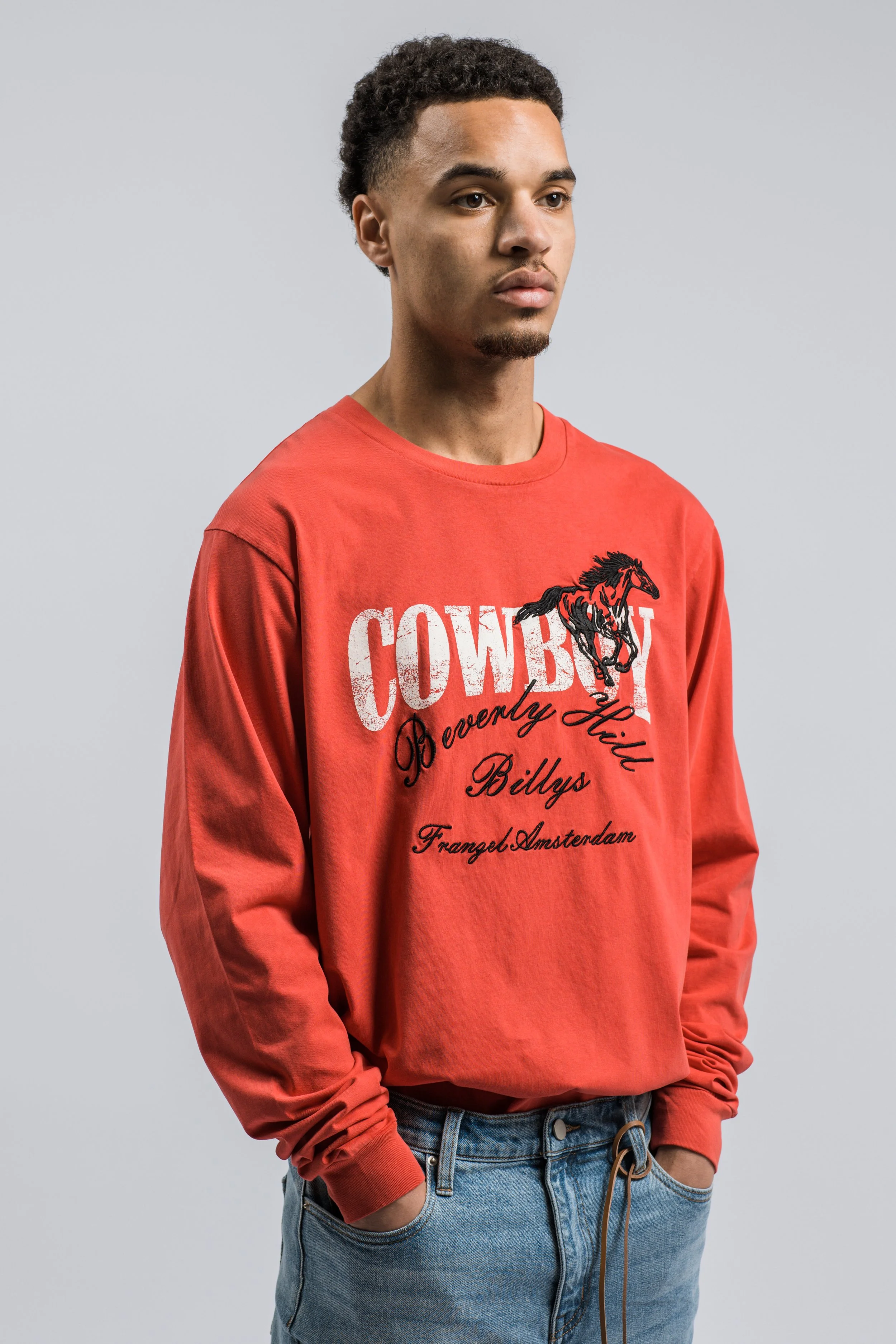 Een jonge man in een rode sweatshirt met een print van een springend paard en tekst, en blauwe spijkerbroek, staat tegen een lichtgrijze achtergrond.