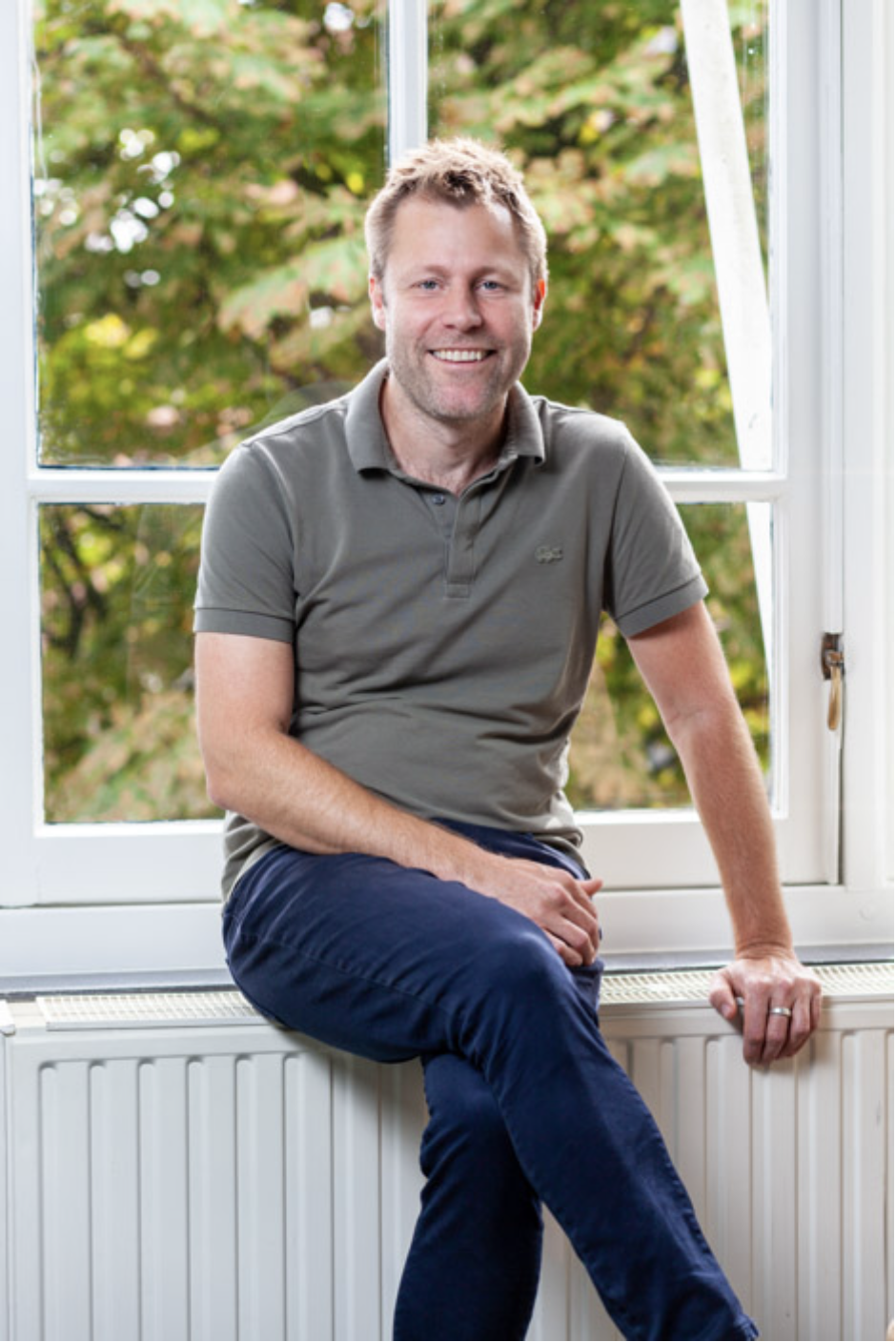 Man zittend op een vensterbank, lachend, met een groene tuin op de achtergrond.