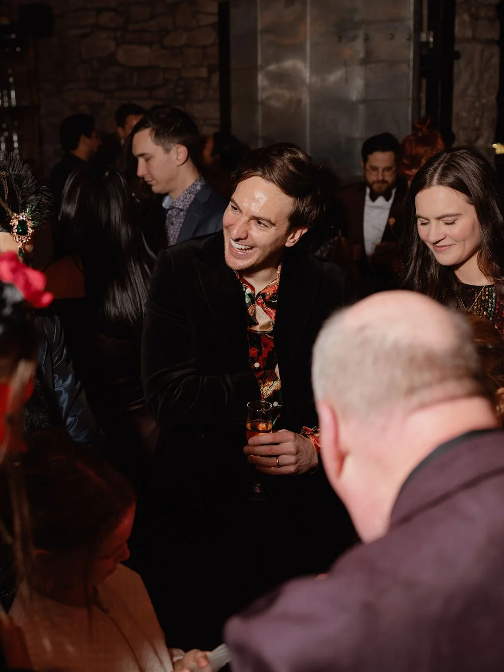 People socializing at a party in a dimly lit venue, with a man smiling and holding a drink in the center, surrounded by others engaged in conversation.