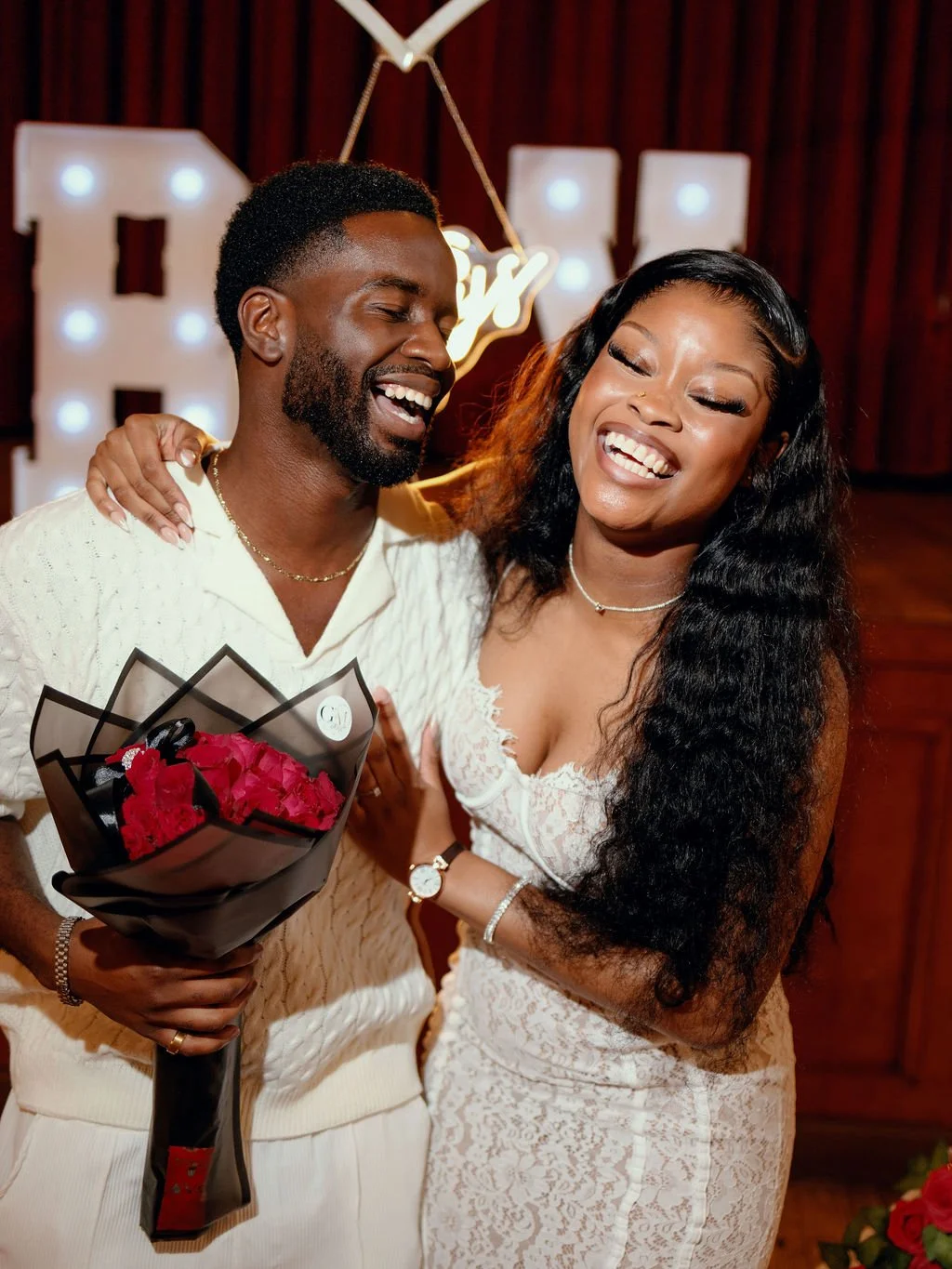 A happy couple at a celebration, with the man holding a bouquet of red flowers, both smiling and dressed in white. The background features illuminated letters and a dark wood wall.