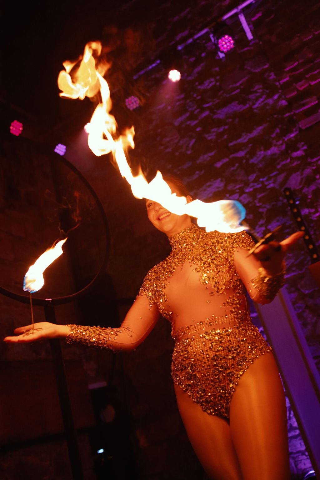 A female performer in a glittering gold outfit is performing a fire act, holding flaming torches in each hand, with flames swirling around her face in a dimly lit venue with purple lighting and a brick wall.