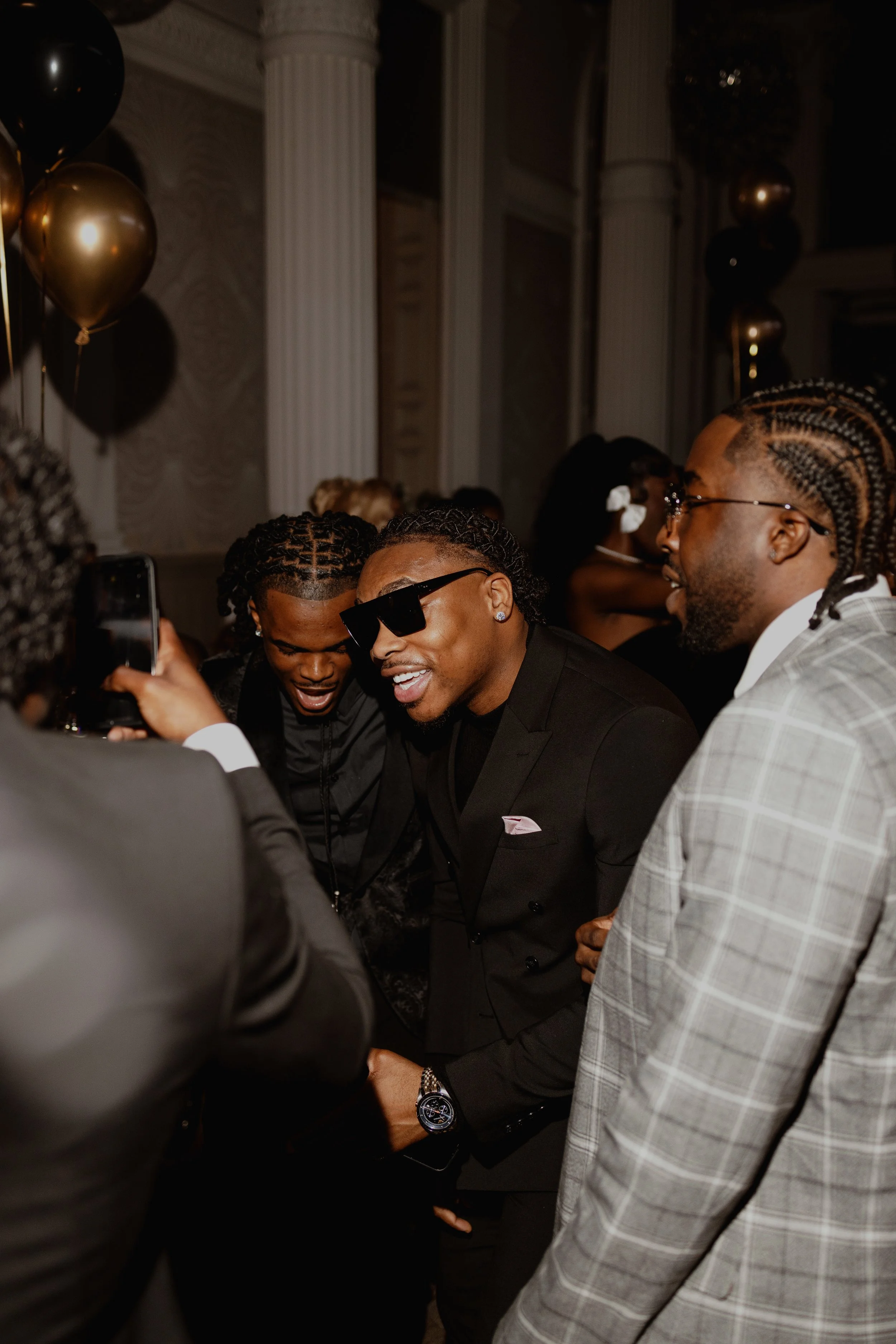 A group of men dressed in formal attire, smiling and taking a selfie at a celebration or party with black and gold balloons and elegant decor in the background.