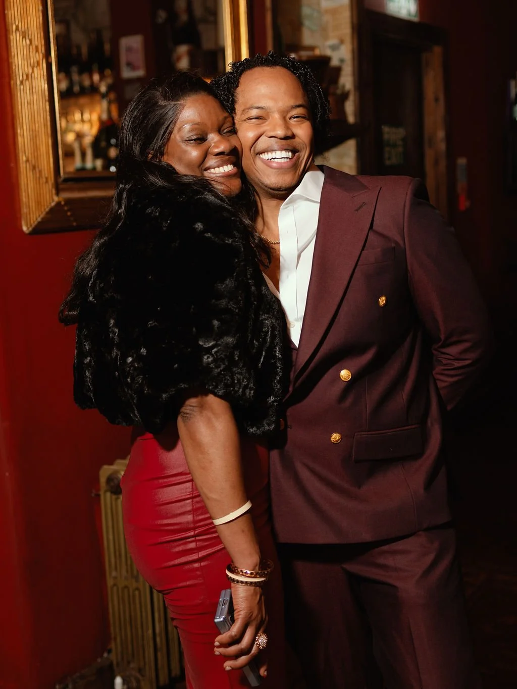 Two people smiling and hugging in a warmly lit bar or restaurant. The woman on the left wears a black fur coat and red pants, and the man on the right is dressed in a maroon suit with a white shirt. They appear happy and enjoying each other's company