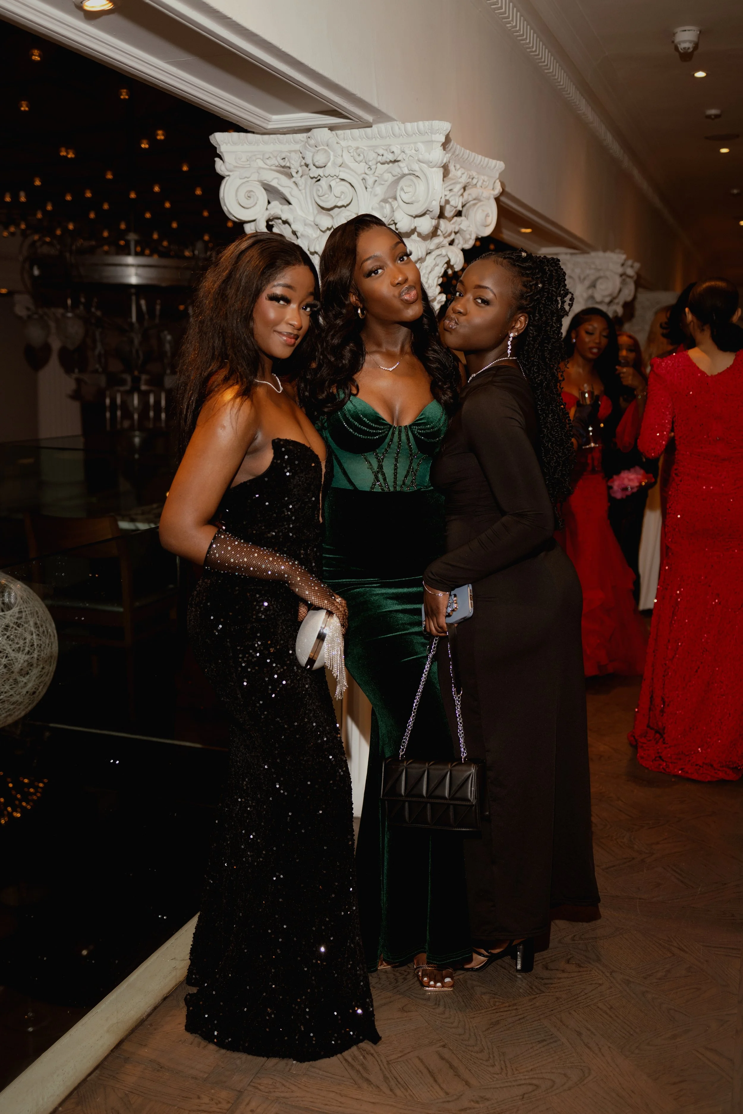 Three women dressed in elegant evening gowns pose together at a formal event, with other guests in the background.