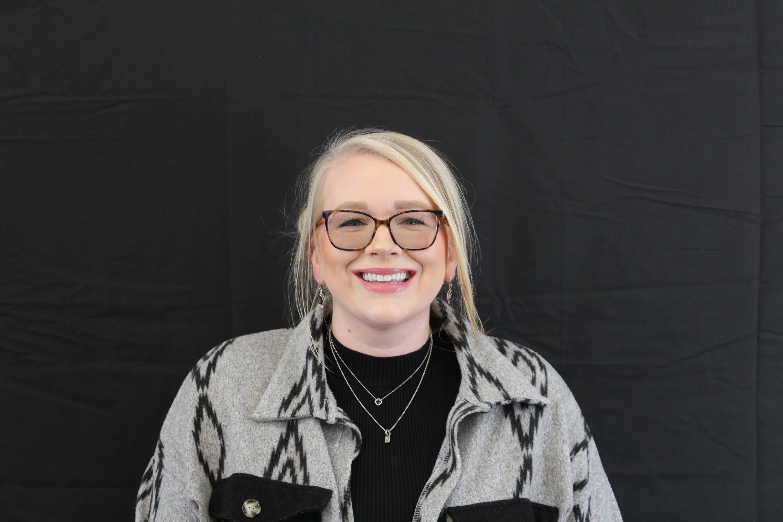 A smiling woman with blonde hair, wearing glasses, earrings, layered necklaces, a black turtleneck, and a gray patterned jacket, standing against a black background.