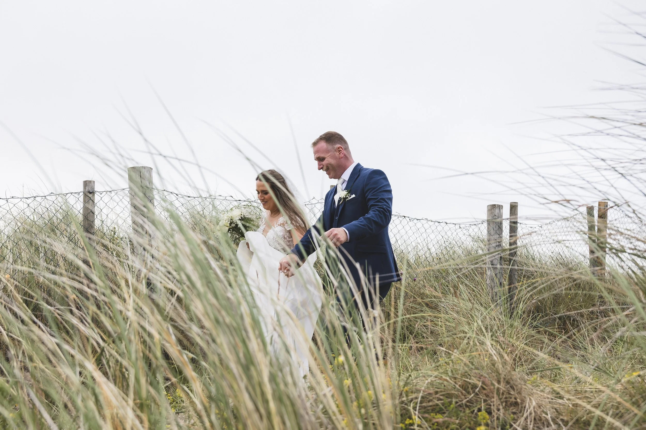 A bride and groom walking through tall grass near a wooden fence, smiling and holding hands during their wedding day outdoors.