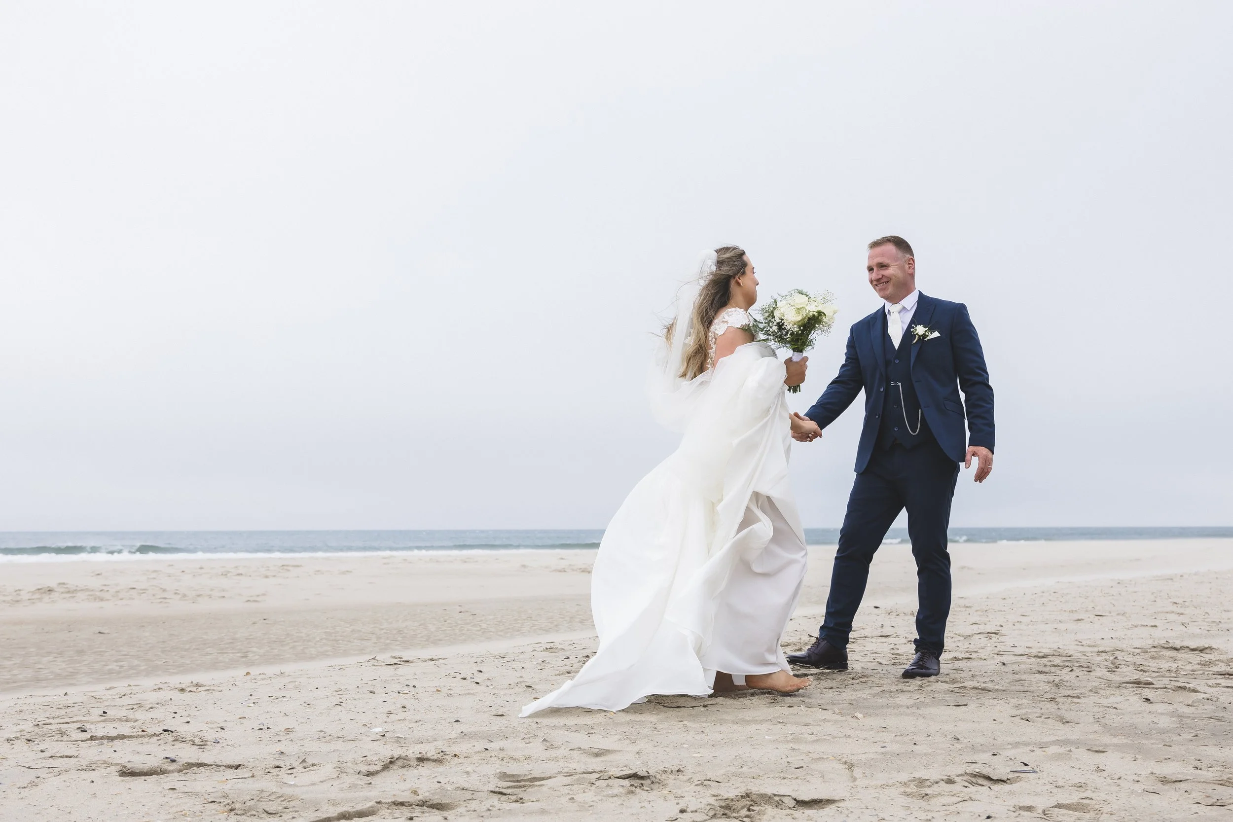Bride and groom holding hands on the beach, with the bride in a wedding dress holding a bouquet of flowers, and the groom in a navy suit, smiling at each other.