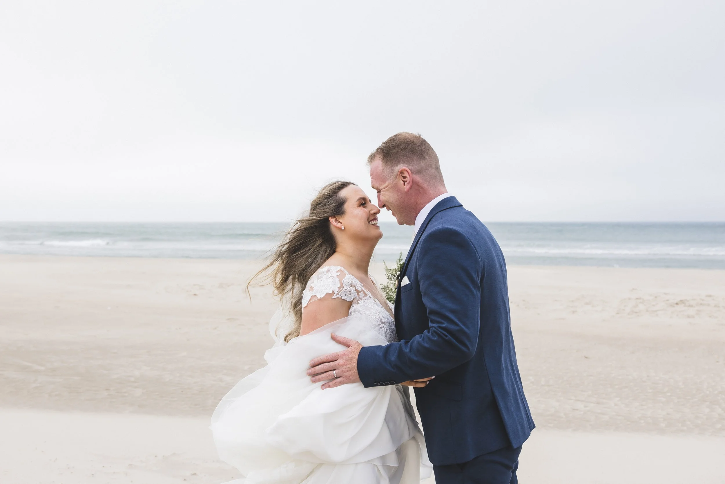 A bride and groom on the beach, smiling and holding each other with waves in the background.