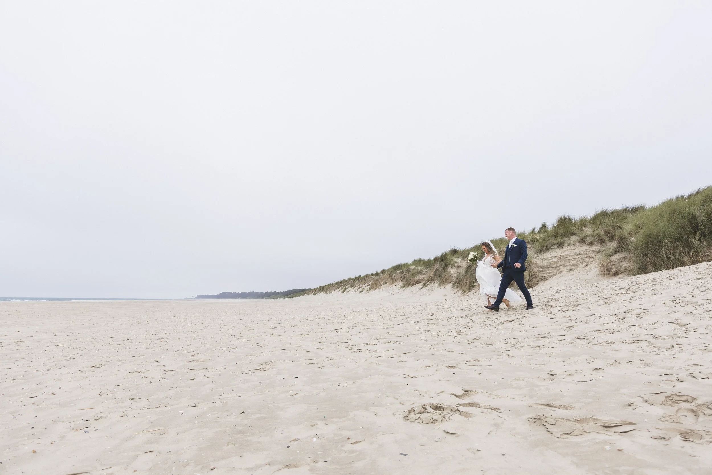 A bride and groom walking on a sandy beach with grassy dunes in the background.