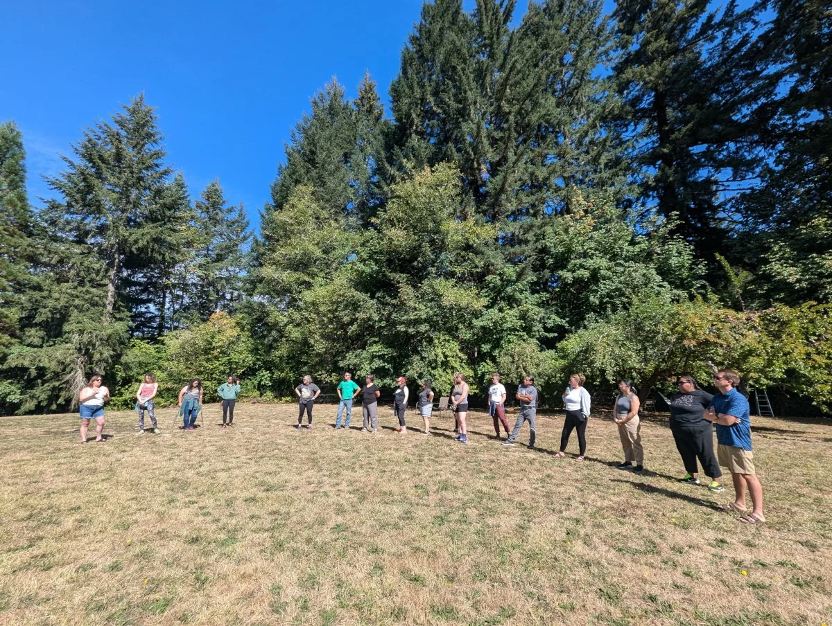 Group of people standing outdoors in a grassy area with tall trees and a bright blue sky in the background.