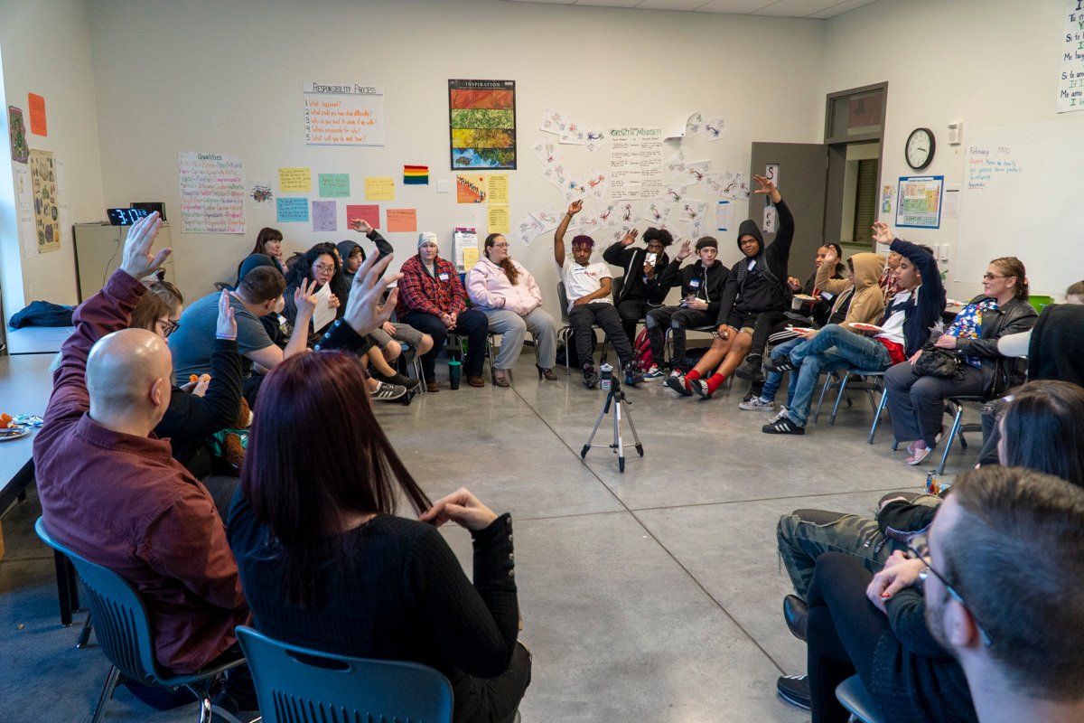 A diverse group of students sitting in a circle in a classroom, some raising their hands during a discussion or activity.
