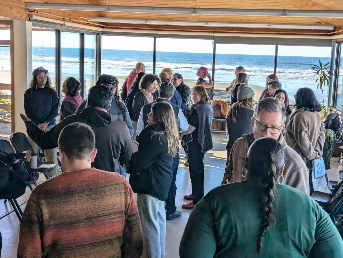 Group of people gathered in a bright room with large windows showing the ocean, some conversing while others wait.