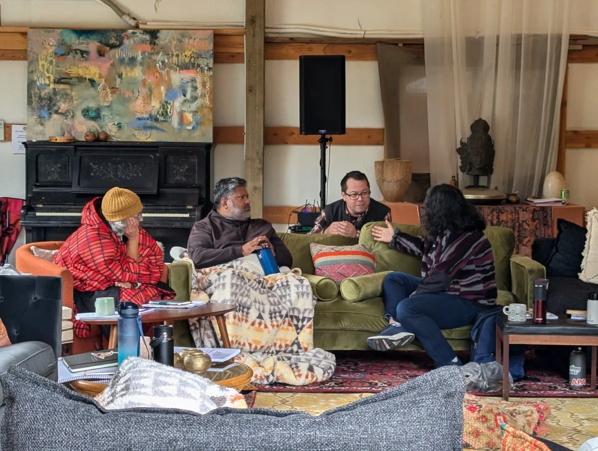 Four people sitting on couches and talking in a cozy, decorated living room with an abstract painting, piano, and various decorative items.