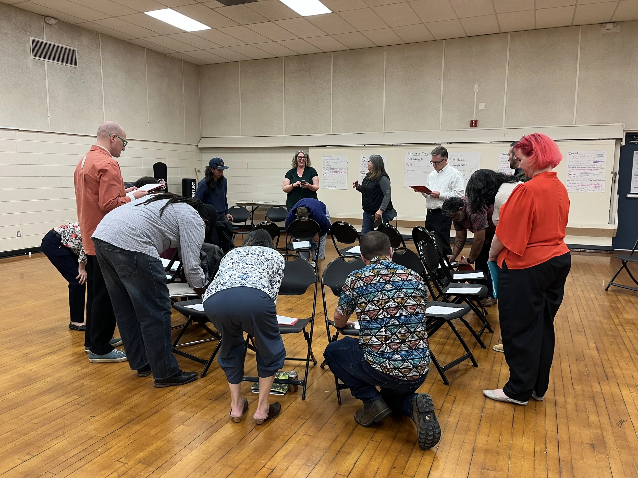 Group of people gathered in a circle in a large room, bowing their heads in prayer or reflection, with chairs and note papers around.
