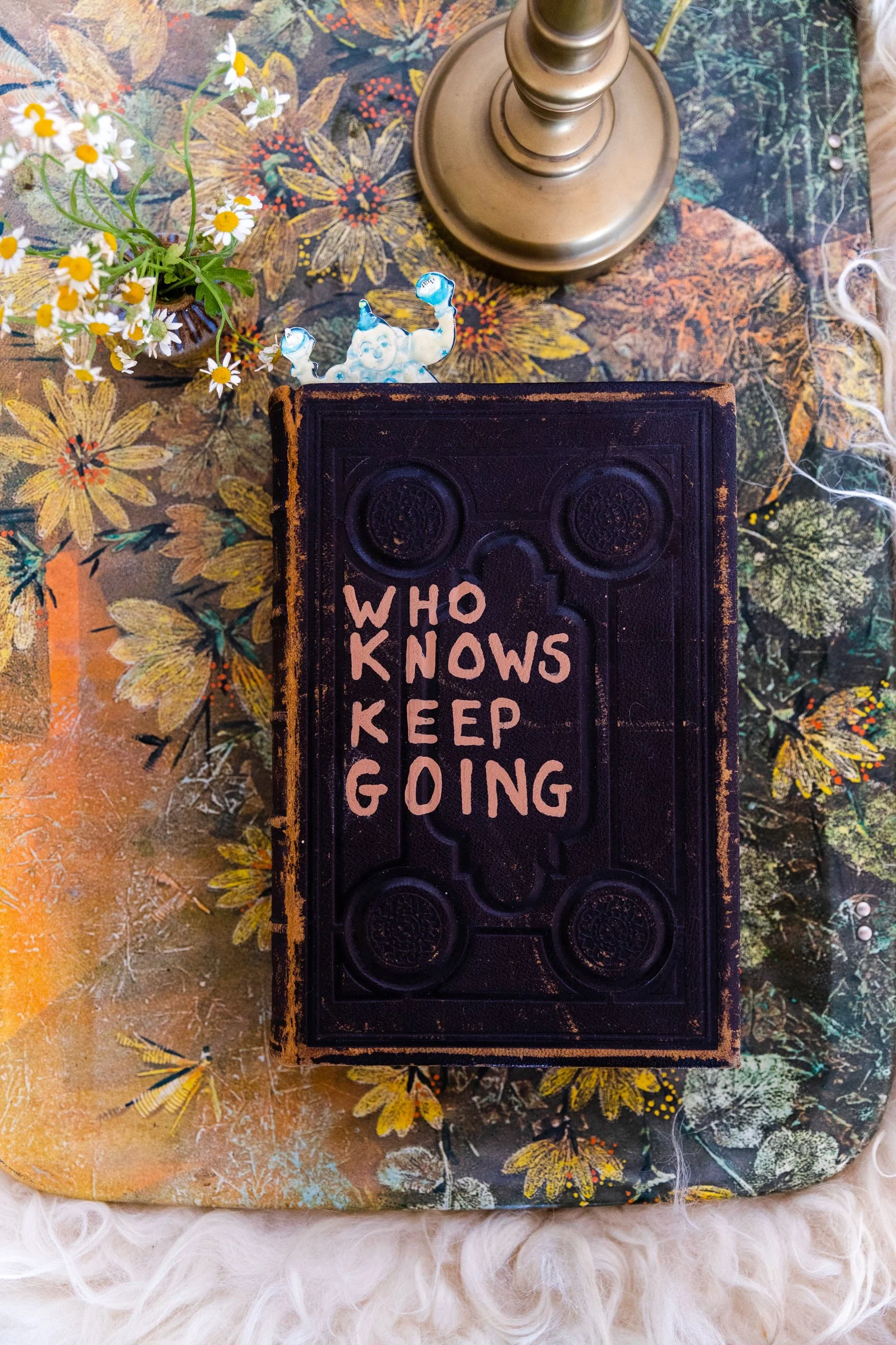 A book with the words "Who Knows Keep Going" written on the cover, placed on a floral table. A small blue and white figurine of a smiling character is on top of the book. There is a brass table lamp and a small bouquet of daisies in a vase nearby.