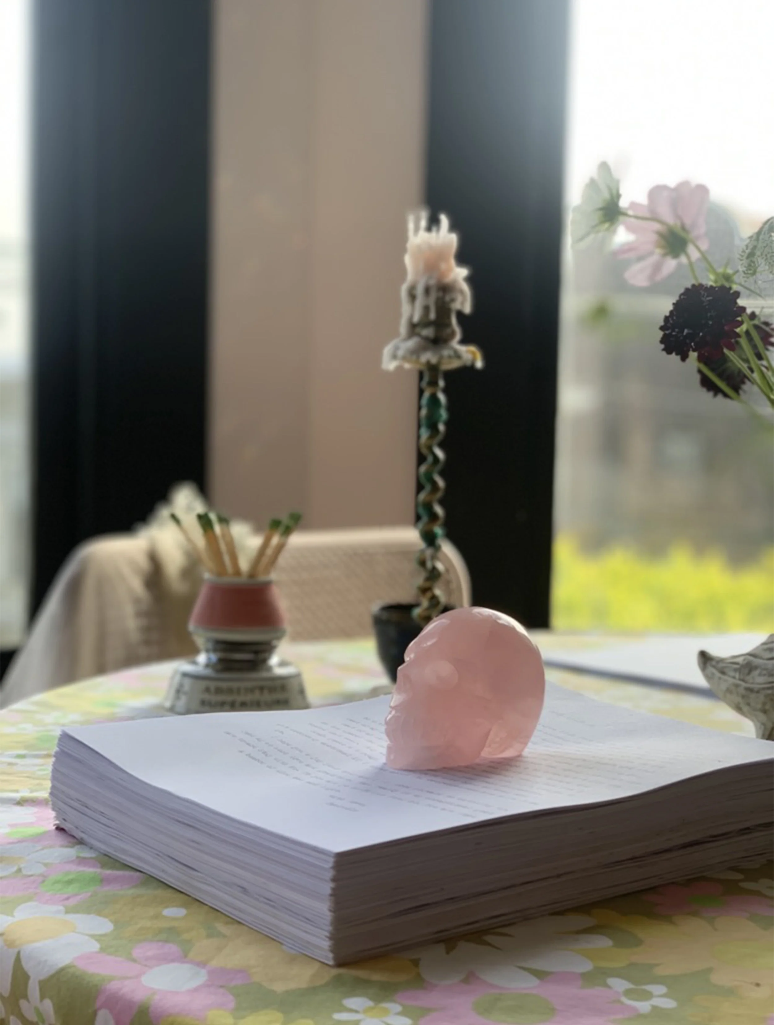 A pink skull sculpture resting on a large pile of papers on a floral tablecloth, with a candle, a candle holder, and a potted plant with flowers in the background.