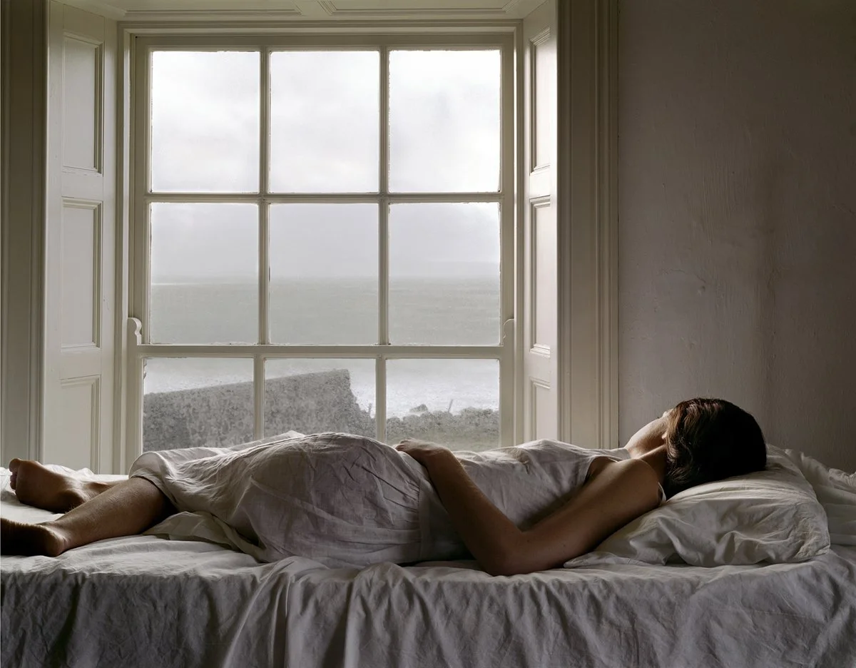 A woman lying on a bed in a room with a large window overlooking the ocean, wearing a white dress and resting on her side.
